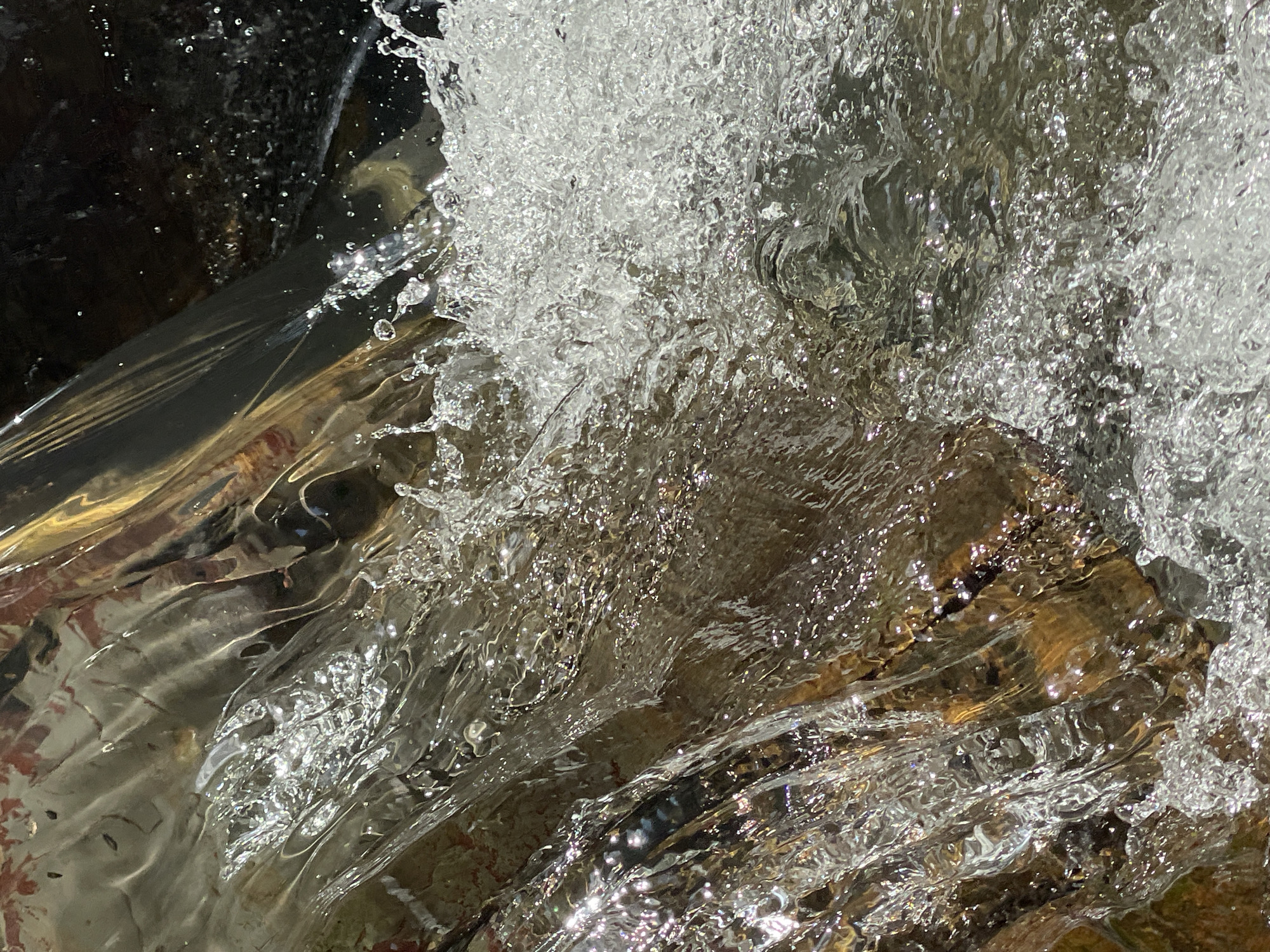 This media file depicts a close-up, dynamic view of a body of water, likely a stream or river, in motion. The primary subject is the water itself, characterized by its turbulence, foaming, and the play of light on its surface. Splashes and ripples are prominent, creating a sense of movement and energy. Beneath the churning water, submerged rocks are visible, providing texture and grounding the scene. The lighting suggests it is daytime, with sunlight catching the water, creating bright highlights and reflecting off the surfaces. The overall impression is one of natural beauty and the raw power of water. There are no people, objects, or text visible in the image, and the location context of Izmaylovka, Russia, is not visually discernible within the frame. The focus is entirely on the abstract patterns and textures formed by the flowing water.