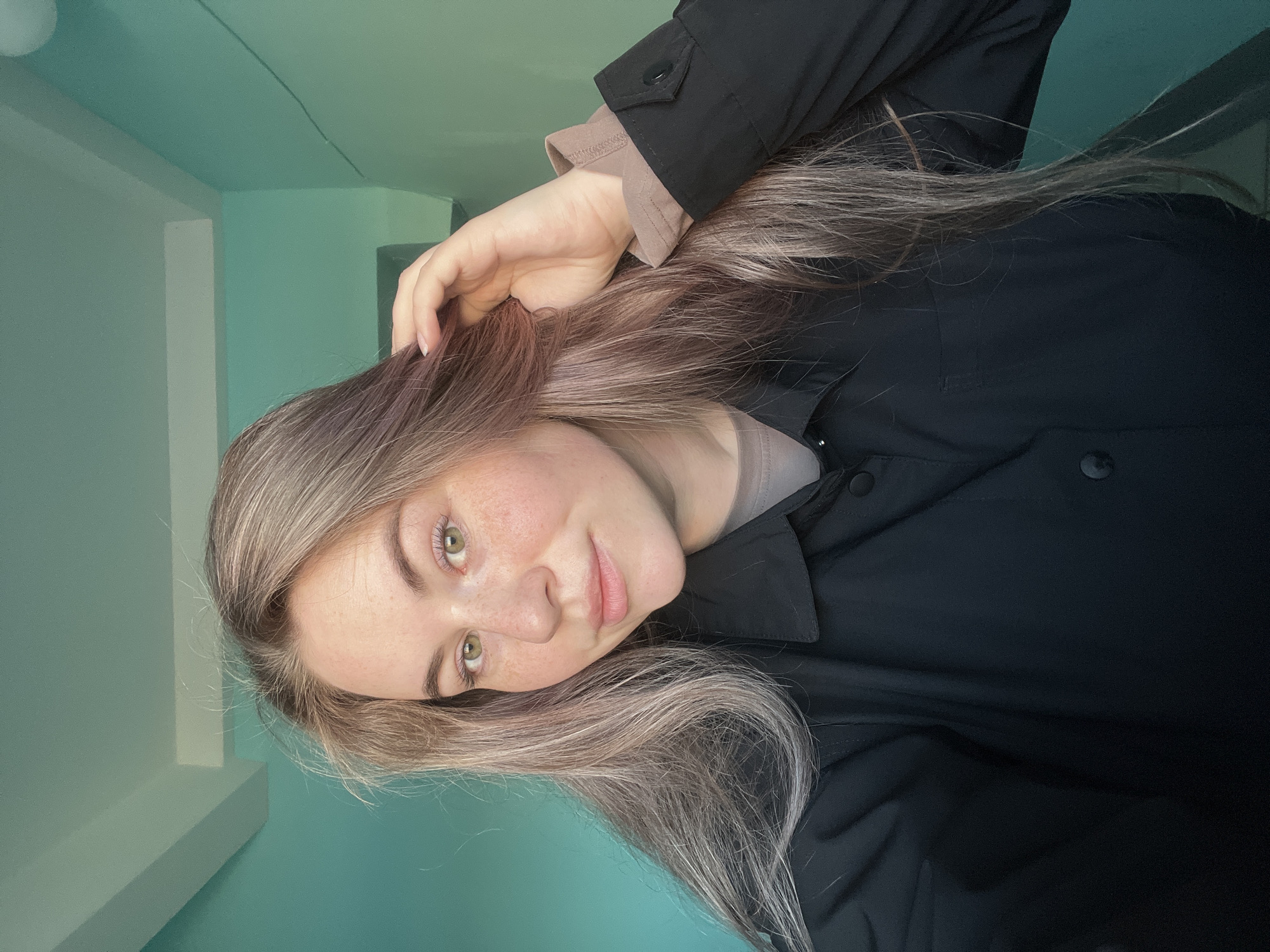 A young woman with fair skin, freckles, and long, light brown and grey hair is posing for a photo. She has light green eyes. She is wearing a black button-down shirt with a beige undershirt. Her right hand is raised to her hair. The background is a pale blue-green wall, possibly indoors. The photo is taken at an angle, with the woman tilting her head slightly. There is no indication of the city of Chaikovsky, Russia, in the image.