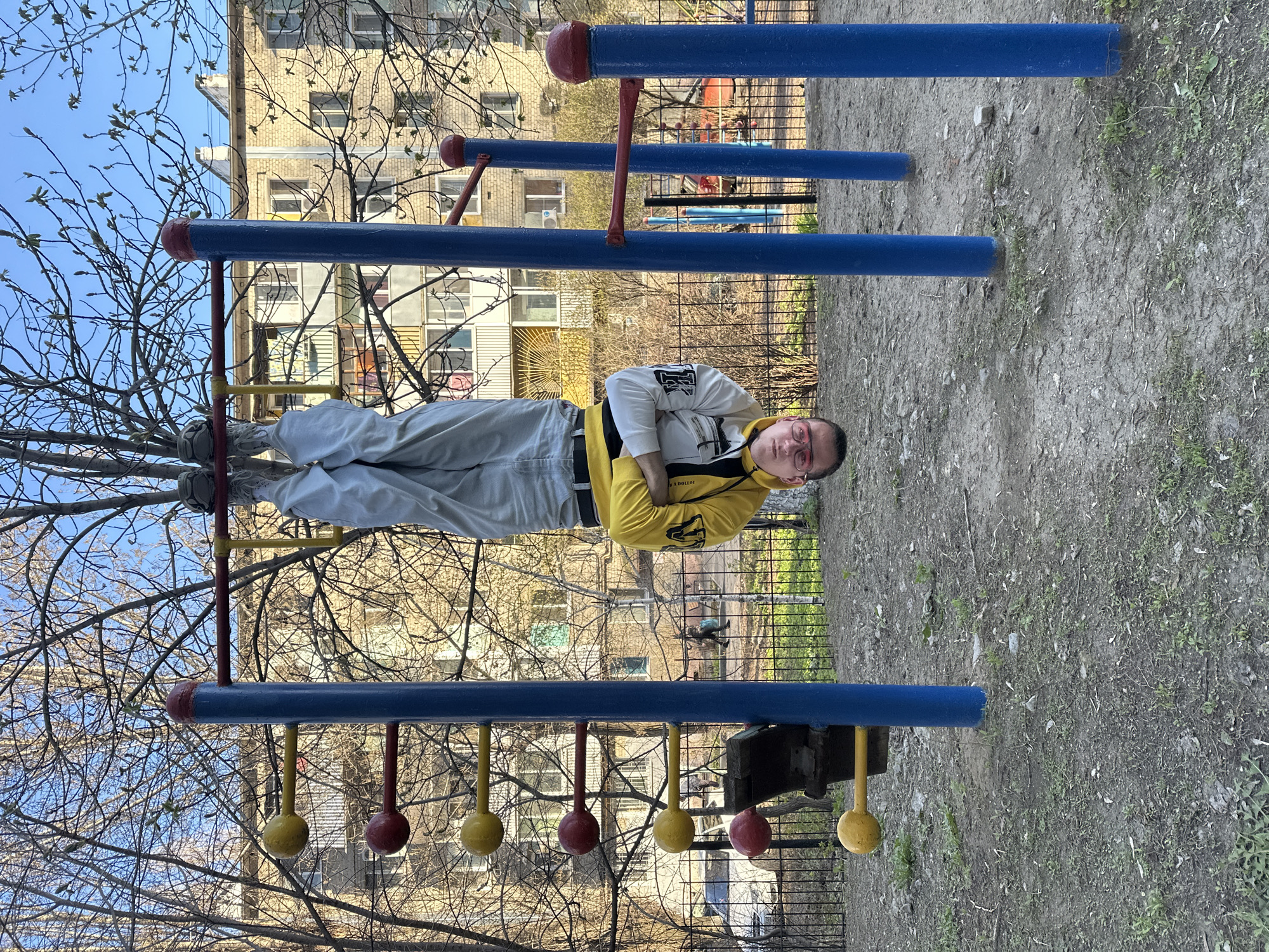The image, taken in Saratov, Russia, captures a person lying horizontally on outdoor playground equipment, creating an intriguing perspective. The scene is set in an outdoor residential area, likely a courtyard, under a bright, clear sky, suggesting daytime in early spring.
The main subject is a young male with light skin and short hair, wearing glasses with reddish frames. He is dressed in a bright yellow hooded sweatshirt over a white long-sleeved top with black graphical details, light grey trousers, and light-colored sneakers. He is positioned on his back, supported by a blue horizontal bar of the playground structure, with his arms crossed over his chest. His expression is neutral, possibly hinting at a playful or relaxed mood.
The playground equipment features prominent blue horizontal bars, red connecting poles, and hanging red and yellow spherical elements, resembling rings or climbing grips. The ground beneath is a mixture of dirt and sparse green vegetation. In the background, typical multi-story brick residential buildings with numerous windows are visible. A black metal fence runs along the perimeter of the area. Bare trees with small, newly budding leaves are scattered throughout the scene, indicating the season.
Visible text on the person's yellow hoodie appears to be "A DOLLOI" or similar, partially obscured, and an indistinct black graphic is on his white undershirt.
