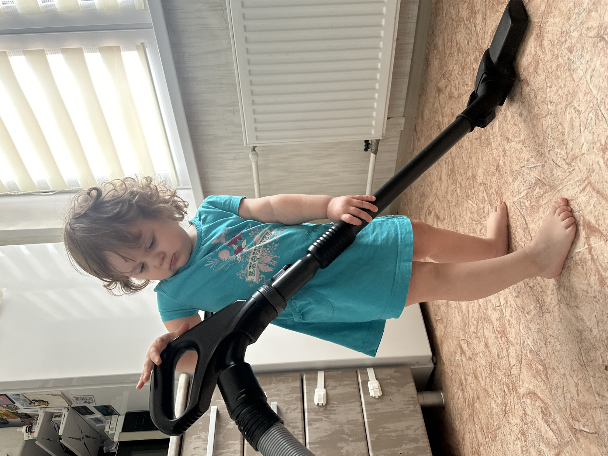 A full-shot image captures a young child, likely a toddler or preschooler, standing barefoot on a light brown, textured laminate or linoleum floor in what appears to be a home in Chaikovsky, Russia. The child has curly brown hair and is wearing a turquoise t-shirt with a floral design and partially visible text, possibly "EKOUTE". The child is holding and seemingly playing with a black vacuum cleaner, gripping the handle of the wand extension with both hands, while the floor head rests on the ground beside their bare feet.
The setting is indoors, brightly lit by natural light streaming through a large white window with light-colored horizontal blinds visible in the upper background. Below the window, a white radiator is mounted on a light-colored, possibly paneled wall. To the child's left, a white cabinet or piece of furniture is visible, with various small items on its surface. To the child's right, part of another wooden-toned cabinet with small white handles can be seen. The child's gaze is directed downwards, focused on the vacuum cleaner or the floor, suggesting a moment of concentration or playful exploration. The overall scene depicts a common domestic moment, with the child engaging in a "helper" activity.