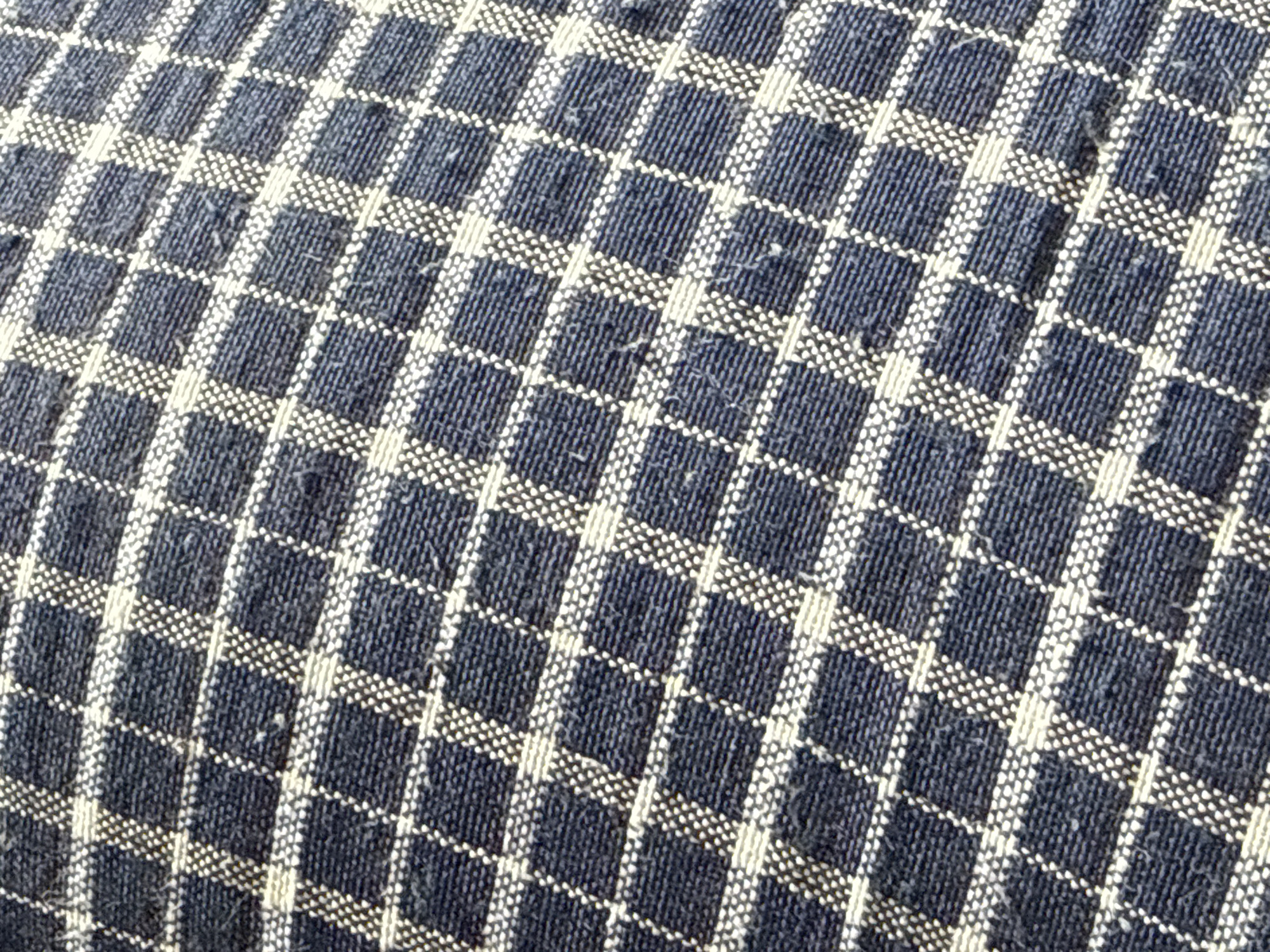 A close-up, high-angle shot captures the detailed texture and pattern of a woven textile. The fabric features a checkered or plaid design, primarily composed of dark blue or indigo squares interspersed with lighter, off-white lines that form a grid. These lighter lines are not solid but appear to be created by numerous small, tightly packed dots or short dashes of light-colored thread, suggesting a particular weave technique or texture.
The dark blue squares also show visible vertical and horizontal thread lines, indicating a coarse or slubby texture within those areas. The overall impression of the fabric is one of a rustic or natural material, possibly cotton or a blend, with a somewhat fuzzy or textured surface. Some very minor loose fibers or slight pilling are visible on the surface, suggesting the fabric might be used or worn.
The fabric itself appears to be draped over a gently curved surface, as the grid lines show a subtle distortion and arc across the frame rather than being perfectly straight. The lighting is soft and even, highlighting the textural details without casting harsh shadows. No specific activity is taking place, nor are there any visible people, objects beyond the fabric, or discernable text. The setting is simply a magnified view of this patterned textile.