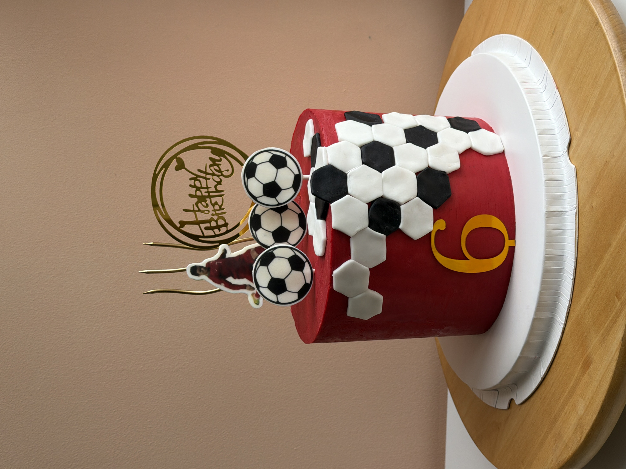 A horizontally-oriented photograph displays a close-up view of a decorated birthday cake. The cake is cylindrical, and covered in red frosting, with a pattern of black and white hexagons resembling a soccer ball on its side. A golden-colored "9" is affixed to the side of the cake. The cake sits on a white circular base, which itself rests on a larger, wooden circular base. Decorations protrude from the top of the cake: a golden "Happy Birthday" topper, three soccer ball decorations on sticks, and a small soccer player figure on a stick. The background is a plain beige wall. The photo does not suggest a location in Tel-Aviv, Israel.