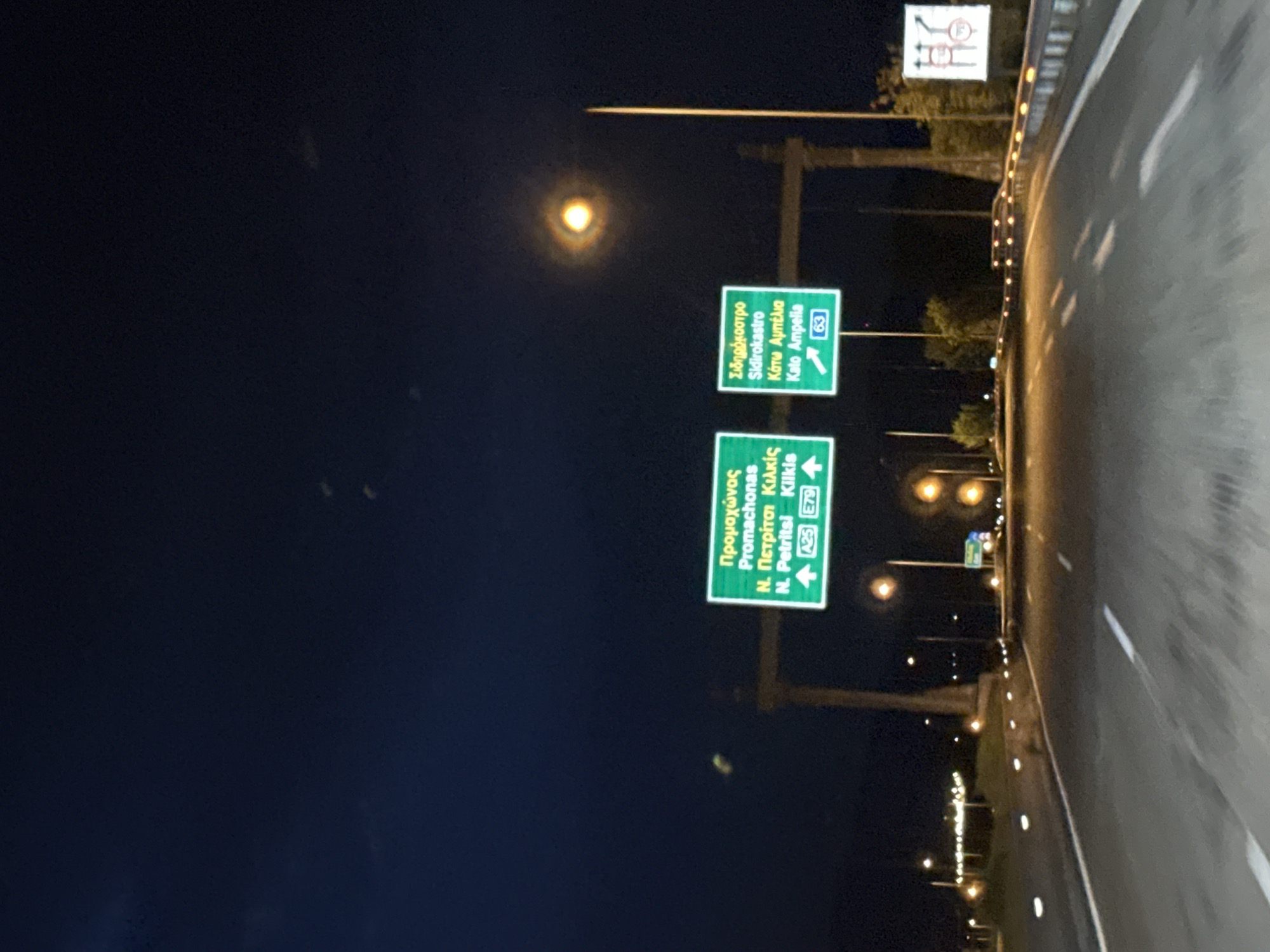 The image shows a road at night in or near Sidirokastro, Greece. Two green highway signs are visible above the road. The top sign indicates directions for Sidirokastro and Kato Ampelia (Route 63). The bottom sign indicates directions for Promachonas, N. Petritsi, and Kilkis (A25 and E79). The road has reflective lane markers. Streetlights are visible along the side of the road.