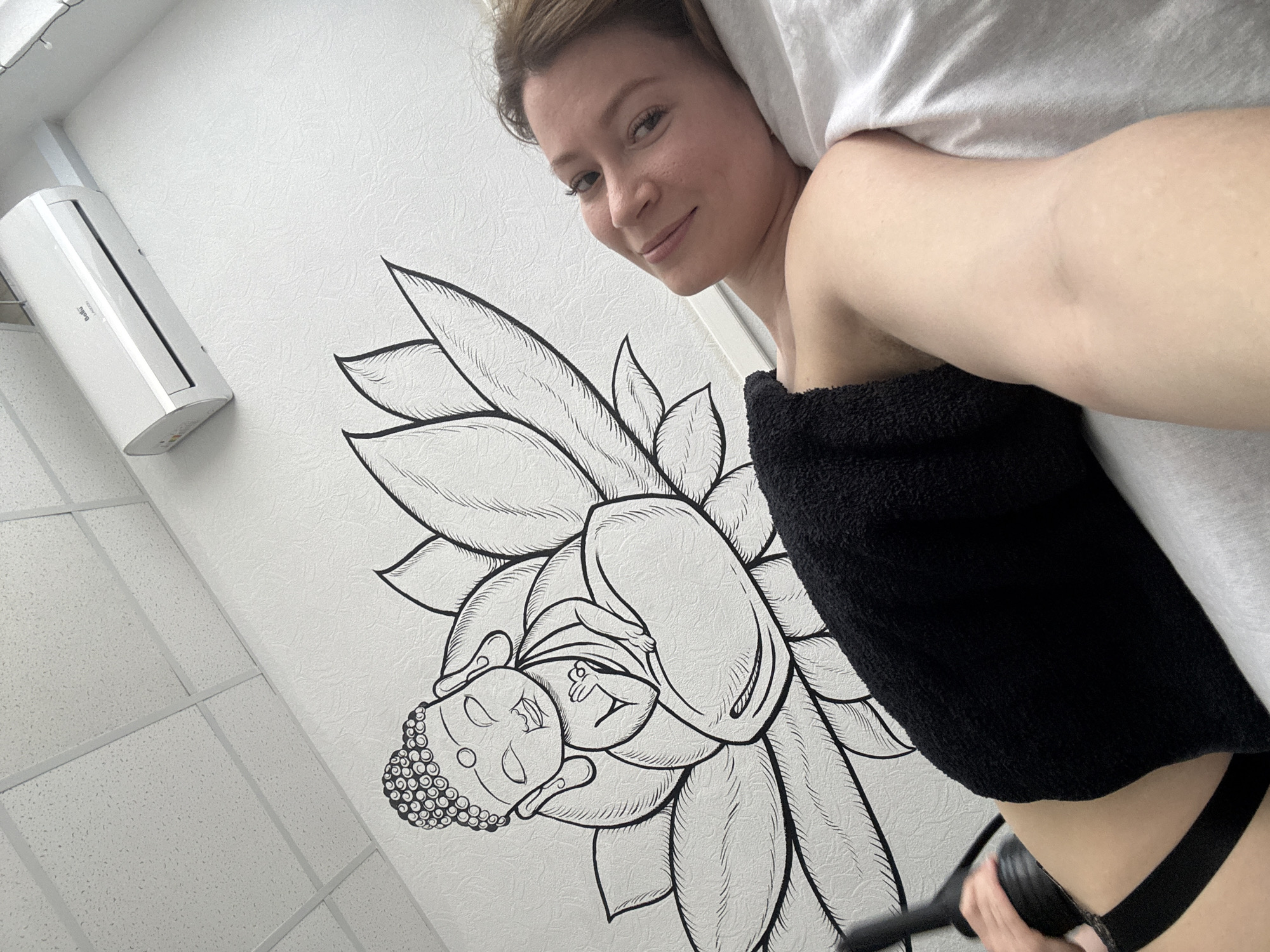 The image shows a woman lying on her side, wrapped in a white sheet and a black towel. She is smiling at the camera. Her lower body is partially visible, with a black strap or belt around her waist. A hand holds a dark-colored device near her hip. The background features a white wall with a black and white drawing of a lotus flower and a Buddha figure. To the left, a white air conditioning unit is mounted on the wall. The scene appears to be indoors, likely in a spa or treatment room. While the location is noted as Chaikovsky, Russia, there are no explicit indicators of this city visible in the image.