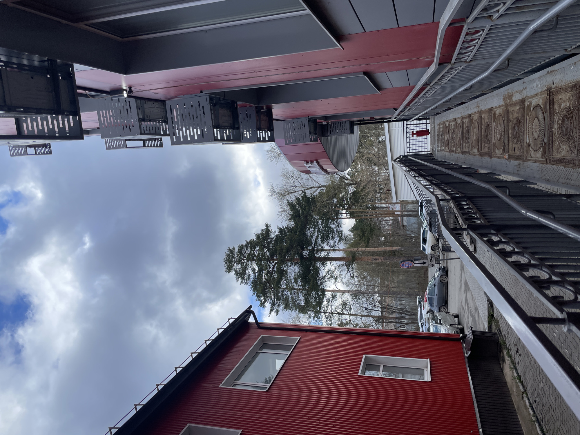 This image captures an outdoor scene in what appears to be a mixed-use or commercial area, with buildings, a walkway, and a parking lot under an overcast sky. The photo is taken from a slightly elevated perspective, looking towards a stand of trees in the distance.
In the foreground, to the left, is a vibrant red building with corrugated metal siding. It features several modern, white-framed windows, including one large, angular window. The building has a dark roofline with a visible downspout.
Above and to the right of the red building, a larger structure is visible, characterized by alternating panels of dark grey and red. Decorative, laser-cut metal panels, featuring a pattern of vertical and horizontal slots, hang from the underside of this building, casting subtle shadows. A light fixture is visible among these panels.
Running from the right side of the image towards the center is a paved walkway or ramp. It is bordered by black metal railings with intricate decorative elements. The surface of the walkway is adorned with multiple rectangular metal inserts, each featuring an embossed circular design.
In the mid-ground, beyond the buildings and walkway, a dense cluster of tall evergreen trees, predominantly pines, stands prominently. Bare deciduous trees are also visible among them, suggesting a season other than full summer. A parking lot is situated to the right, adjacent to the walkway, where several cars are parked, including a silver, a dark grey, and a white vehicle. A red circular "No Entry" road sign, with a white bar and an arrow pointing to the right, is visible near the parked cars. A small red box, possibly a fire extinguisher cabinet, is mounted on a fence further along the walkway.
The sky is heavily overcast with prominent grey and white clouds, though a patch of brighter blue sky is discernible towards the far left. The overall lighting is diffuse, indicative of daytime under cloudy conditions. There are no people visible, and no specific activities are taking place. The location context provided is Izhevsk, Russia.