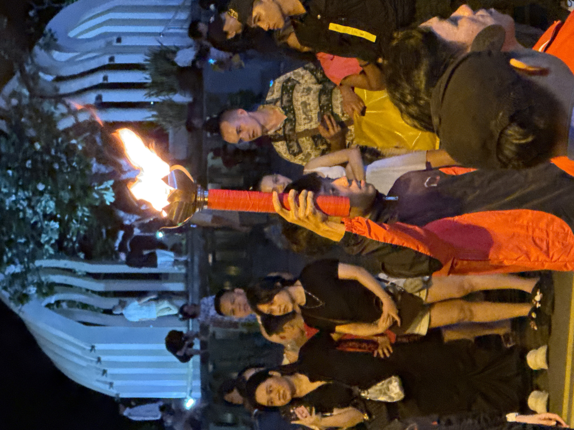 An outdoor night-time crowd is gathered near a large, modern white structure with vertical slats. A person in the foreground is holding a large torch with a bright flame. Several individuals are visible in the midground and background, some observing the torch. The scene appears to be part of a public event or ceremony in Nha Trang, Vietnam.