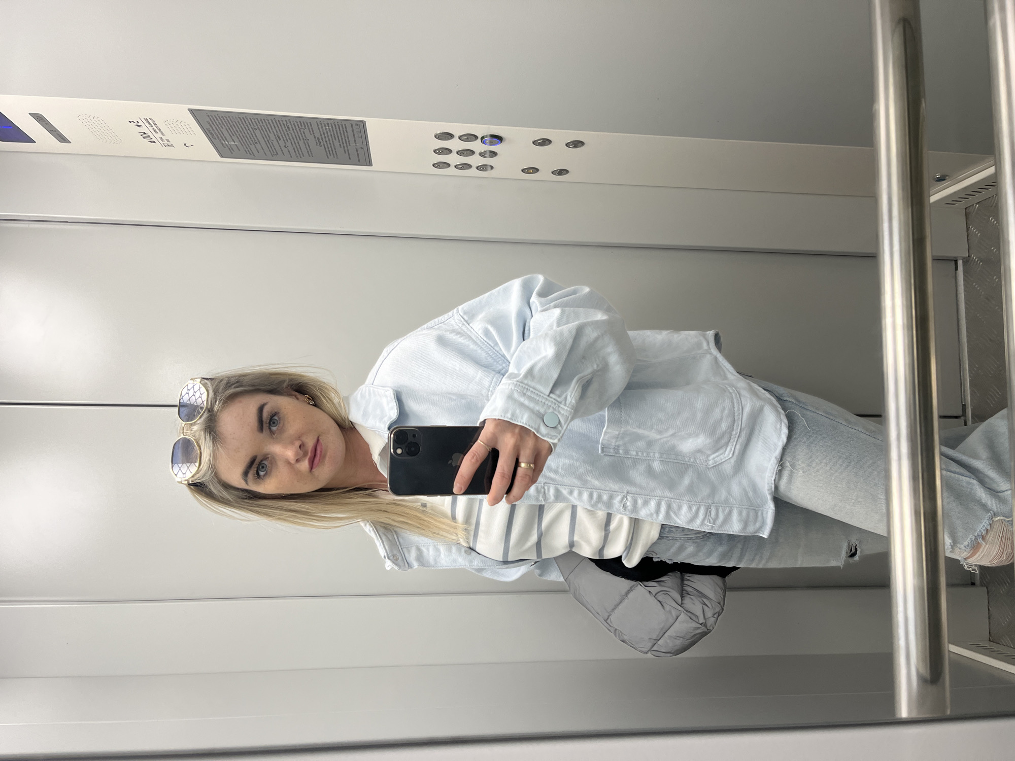 A blonde woman is taking a selfie in an elevator. She is wearing sunglasses on her head, a light blue denim jacket, a striped shirt, and light-wash ripped jeans. She is holding a black smartphone in her right hand. The elevator is modern and has stainless steel handrails, a control panel with illuminated buttons, and informational panels. The location appears to be inside a building in Chaikovsky, Russia, based on the presence of Cyrillic text on the elevator control panel.