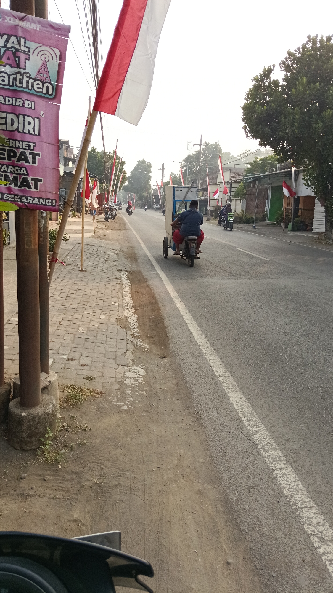The image depicts a street scene in Ploso, Indonesia, likely during a festive or commemorative occasion, indicated by numerous Indonesian flags displayed along the road. In the foreground, a pink banner promoting "XL SMART" and "Smartfren" with services like "INTERNET CEPAT" (fast internet) and "HARGA HEMAT" (economical price) is visible, attached to metal poles. To the right of the banner, a paved walkway lines the road, which is made of asphalt with a white dividing line.
The road itself shows signs of light traffic. A man wearing a blue shirt and red pants is riding a motorized tricycle cart, moving away from the camera. The cart appears to be a food or goods vendor. Further down the road, several motorcycles are parked along the side and some are in motion. A few other individuals are visible on motorcycles, also moving in the same direction. The background shows buildings, trees, and a generally bright, overcast sky. The overall atmosphere suggests a normal day with some local embellishments.