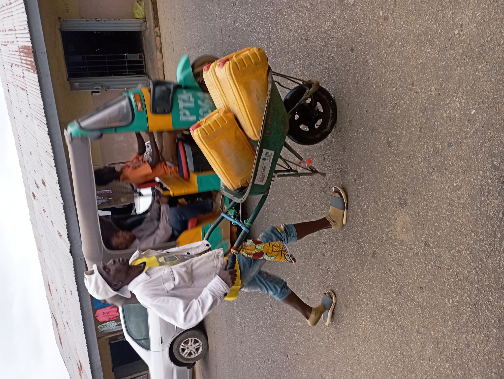 In Barakin Akawo, Nigeria, a man walks down a paved road, pushing a wheelbarrow laden with three yellow jerrycans. He is wearing a white baseball cap, a light-colored jacket over a yellow shirt, blue denim shorts, and sandals. The wheelbarrow has a dark green basin and a single wheel. Behind the man, a three-wheeled vehicle, possibly a tricycle or tuk-tuk, with a driver wearing an orange shirt and a passenger, drives away. Buildings with corrugated metal roofs line the street, with one building appearing to be a shop with a metal gate. A silver car is parked on the left side of the road. The sky is overcast.