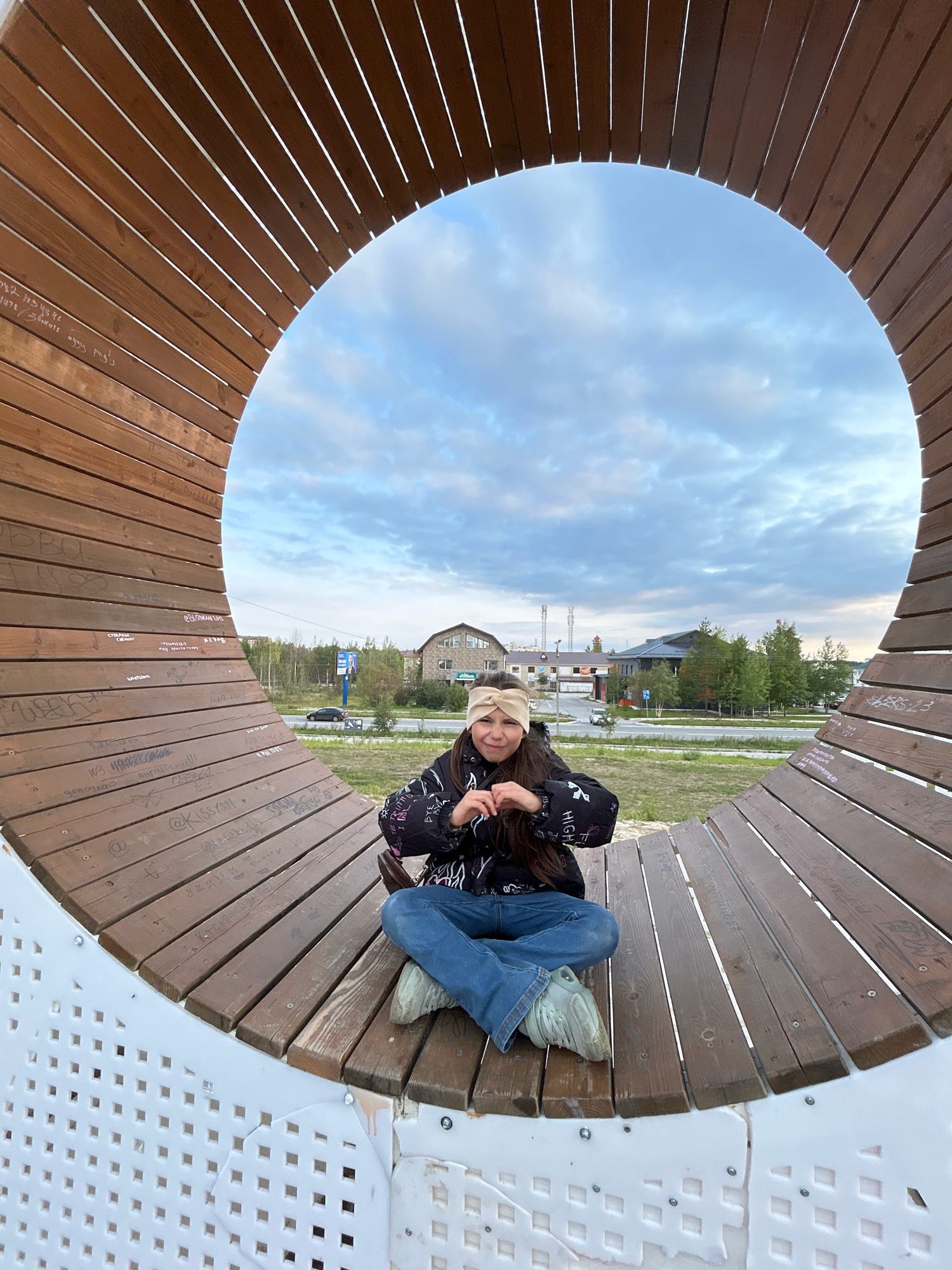 In Noyabrsk, Russia, a young woman is seated cross-legged on a wooden structure, which appears to be a bench or seating area, within a large, circular wooden frame. The frame itself is constructed from curved wooden planks, forming a tunnel-like opening. The woman is wearing a light-colored headband, a dark puffer jacket with white patterns, and blue jeans. She is looking at the camera and has her hands forming a heart shape near her chest.
The wooden surface she is sitting on is covered in graffiti and inscriptions. The background visible through the circular opening shows a cloudy sky with a hint of blue. Beyond that, there are buildings, trees, a road with a car, and industrial structures with tall towers in the distance, suggesting an urban or semi-urban setting. The wooden structure has a white, perforated base. The overall scene captures a moment of leisure in an outdoor public space.