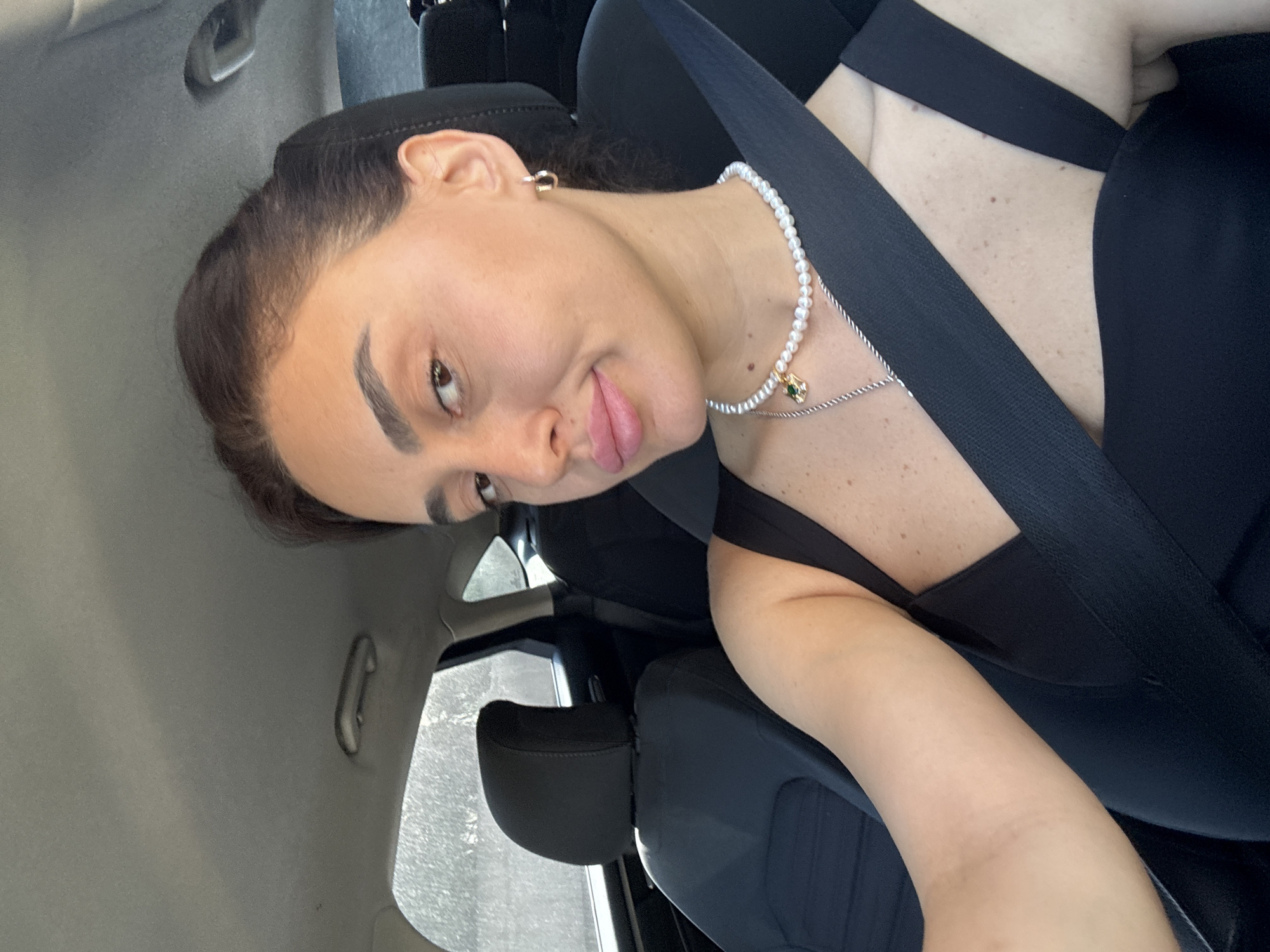 A young woman with dark hair pulled back is in the passenger seat of a car, looking at the camera. She is wearing a black top, a pearl necklace with a green pendant, and small hoop earrings. A seatbelt is across her chest. The car interior is visible, with dark seats and a gray headliner. Sunlight streams in from the left, illuminating the interior and casting shadows. The background outside the car window is blurred, but suggests trees and foliage. The image appears to be taken during the daytime. The location is not specified as Tbilisi, Georgia.