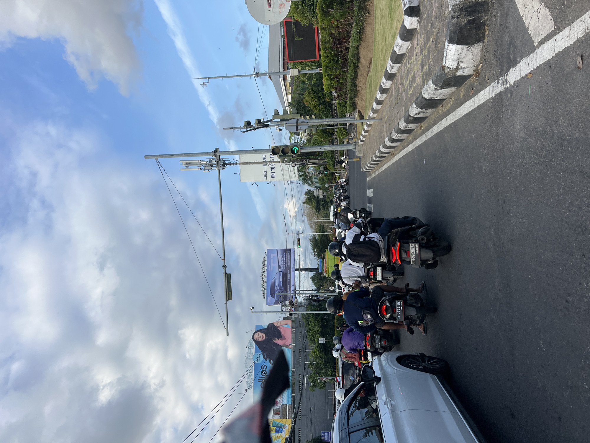 A street scene in Denpasar Barat, Indonesia, features a busy road with multiple lanes of traffic. A white car is positioned in the leftmost lane, facing forward. Behind it, a long line of motorcycles and scooters navigates the road, with riders wearing helmets. In the background, large billboards are visible, one displaying a woman and text that reads "ellips". Another billboard shows a car and the text "BAIC T1 GET READY TO EXPLORE!". Traffic lights, some displaying green arrows, are positioned above the road. Utility poles with cellular antennas are also present. The sky is partly cloudy with patches of blue visible. The road is lined with a curb painted in black and white stripes, and a strip of green grass borders the curb.