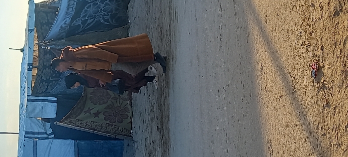 The image depicts two individuals walking from left to right across a dusty, sandy foreground in Khan Yunis, Palestinian Territory. In the background, a cluster of makeshift shelters is visible, constructed from blue and white tarpaulins, as well as patterned blankets or rugs, one featuring a floral and star motif.
The individual in the foreground, a male, wears a long, light brown hooded overcoat over a mustard yellow shirt and light grey trousers. He has short, dark hair and is looking towards his right. Behind him, a female individual wears a dark headscarf (hijab) and a long, dark brown dress or abaya. She carries a dark plastic bag in her left hand and wears white sneakers with dark stripes on her feet. Both individuals are in mid-stride.
Long shadows are cast on the ground from an unseen light source, suggesting either early morning or late afternoon. In the lower right foreground, a small, circular red and metallic object lies on the sand. The ground is uneven, characterized by packed dirt and sand.