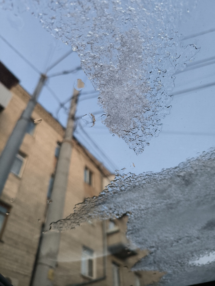 The image shows a close-up view through a car windshield covered with melting ice. The ice has a textured, crystalline appearance, with some clear patches revealing a bright, overcast sky. Behind the windshield and out of focus is a multi-story building with tan brick and several windows. Also visible are utility poles and electrical wires, adding to the urban backdrop. The ice obscures the view, creating a distorted and fragmented perspective.