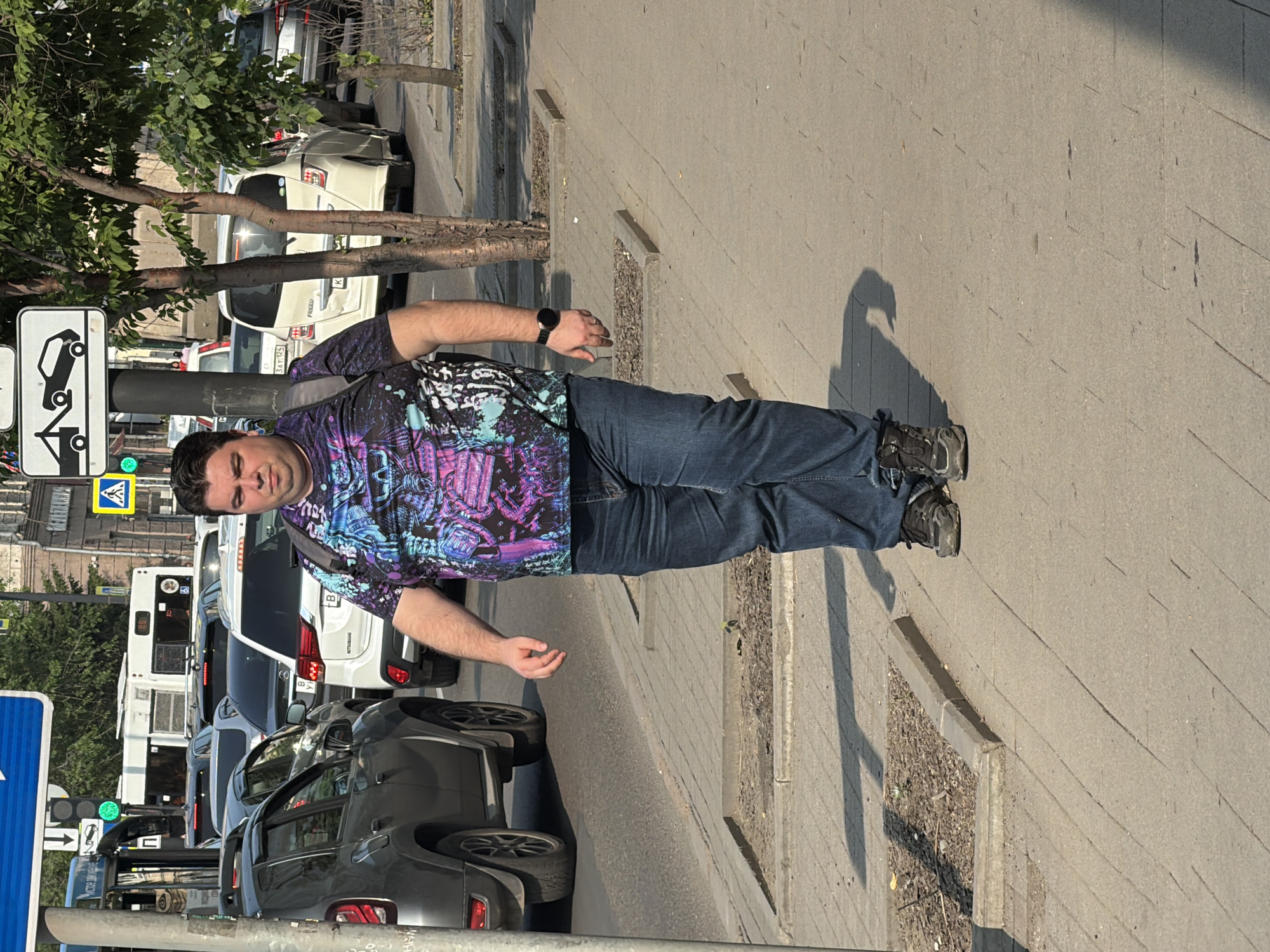 A man is walking on a sidewalk in Krasnoyarsk, Russia. He is wearing a dark t-shirt with a colorful graphic print, blue jeans, and dark athletic shoes. He has a backpack on his shoulders and a watch on his left wrist. He is walking towards the right of the frame. In the background, there is a street with traffic, including cars and a bus. There are also trees and buildings visible. The weather appears to be sunny.