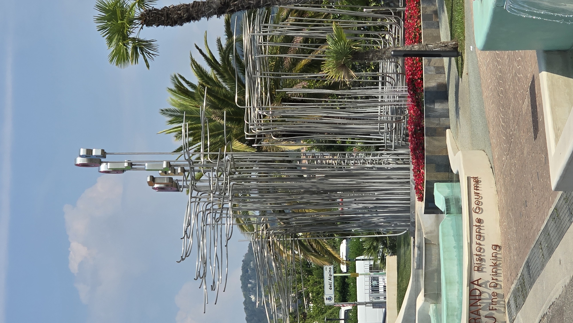This is a photograph of a public art installation in Bardolino, Italy. The installation features a complex structure made of interconnected metal tubes, with several circular elements at the top that appear to be made of chrome and colored glass or plastic. The sculpture is situated in front of a backdrop of palm trees and lush greenery, with a hillside visible in the distance. In the foreground, there is a modern fountain with turquoise water, surrounded by curved concrete structures. Text is visible on one of the concrete structures, reading "GRANDA Ristorante Gourmet" and "Fine Drinking". A street sign indicating "via dell'Alpino" is also present, suggesting the installation is located near that street. The sky is blue with some scattered clouds. The scene appears to be a public space, likely a plaza or park, designed for leisure and dining, given the presence of the restaurant signage and fountain. There are no people visible in the image.