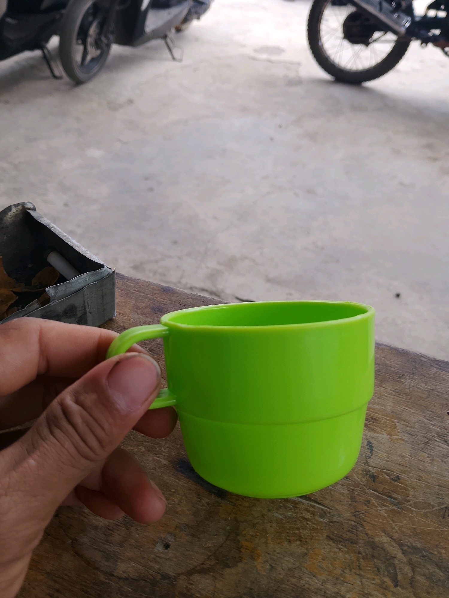 In the city of Unknown, Unknown, a person's hand, with visible fingernails and creases, holds a bright green plastic mug. The mug is positioned on a weathered wooden surface. To the left of the mug, a metallic object, possibly an ashtray, contains what appears to be cigarette butts and paper. In the background, out of focus, the wheels and parts of two motorcycles are visible. The ground beneath the motorcycles is a concrete surface.