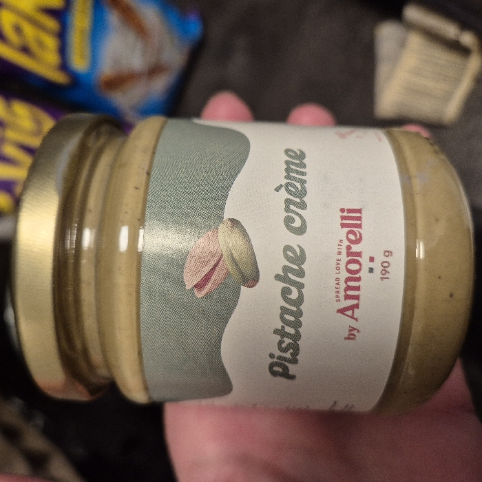 A glass jar of "Pistache crème by Amorelli," weighing 190g, is held upright by a person's left hand. The jar features a gold-colored metallic lid and contains a pale green/beige spread. Its white label is adorned with the text "Pistache crème" in a dark green, slightly cursive font, overlaid on a light green textured background with a wavy white border. An illustration of two pistachios, one partially open revealing a pinkish-red kernel, the other closed and green-brown, is centered on the label. Below, the brand "Amorelli" and the tagline "SPREAD LOVE with" are printed in red, alongside the weight "190 g" and a small red square symbol. The background is out of focus, revealing a blurred blue and purple snack bag in the upper right quadrant and a crumpled light-colored object, possibly a paper cupcake liner, in the lower right. The scene is indoors under artificial light. This item is located in the city of Strasbourg, France.