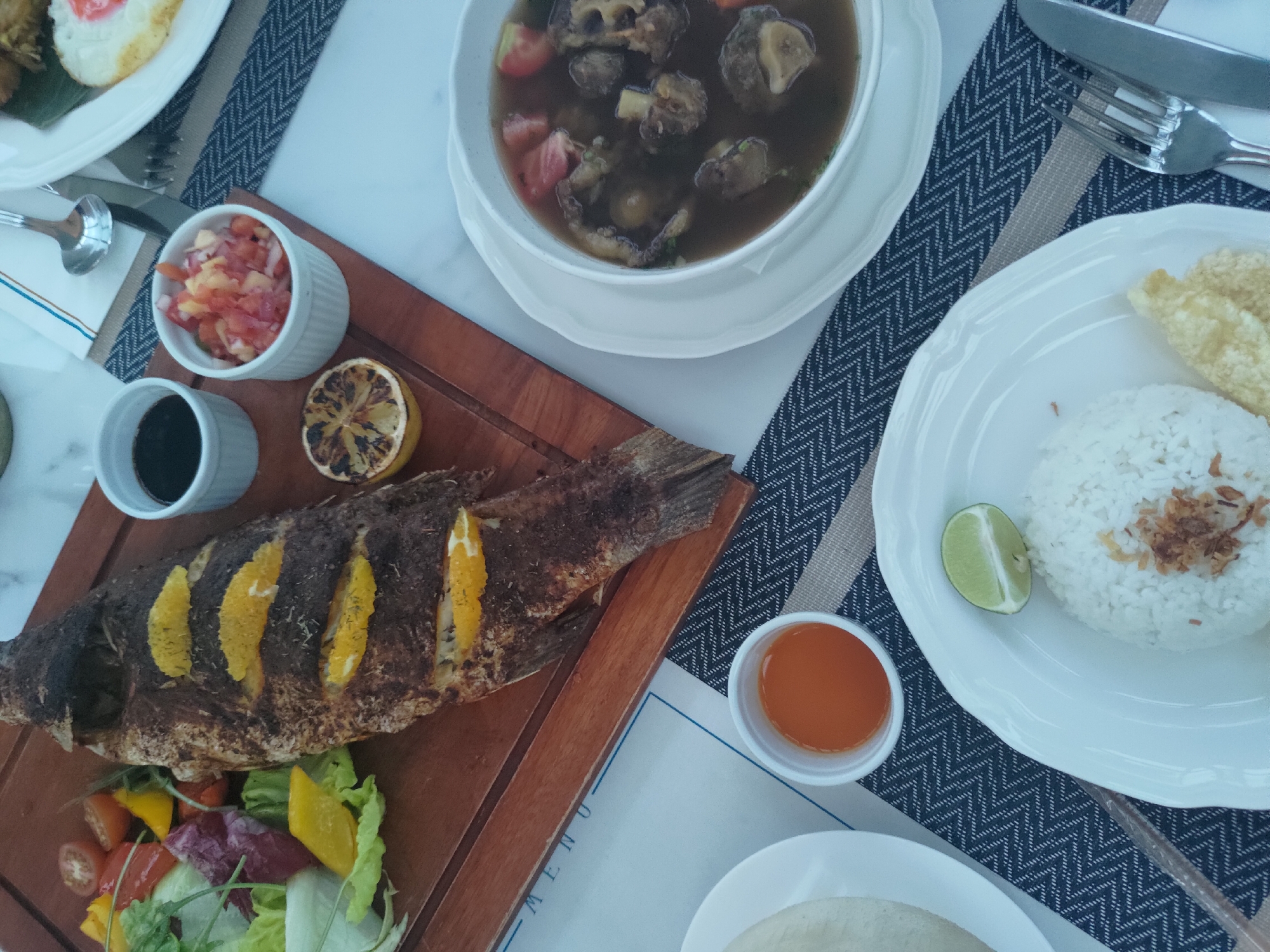 The image shows a close-up, top-down view of a table setting with a meal. A whole grilled fish, garnished with orange slices, is presented on a wooden board alongside a fresh salad composed of lettuce, cherry tomatoes, and bell peppers. There are three small white bowls: one containing a dark liquid, likely soy sauce; another with a finely diced red and white mixture, possibly a salsa or relish; and a third with an orange sauce. A grilled lemon slice is also visible on the board.
To the right of the board, a white bowl holds a dark, savory broth with chunks of meat and tomatoes, suggesting a soup or stew. A plate of white rice, topped with fried shallots and a lime wedge, sits in the foreground. To the left of the rice, a white, rounded object, possibly a steamed bun or bread, is partially visible on a white plate. A fork and knife are placed to the right of the rice plate. The table is covered with a dark blue and white patterned placemat and a beige runner. The background suggests an indoor dining area, with hints of a marble tabletop and a menu. The overall presentation suggests a restaurant setting in Kosambi Barat, Indonesia.