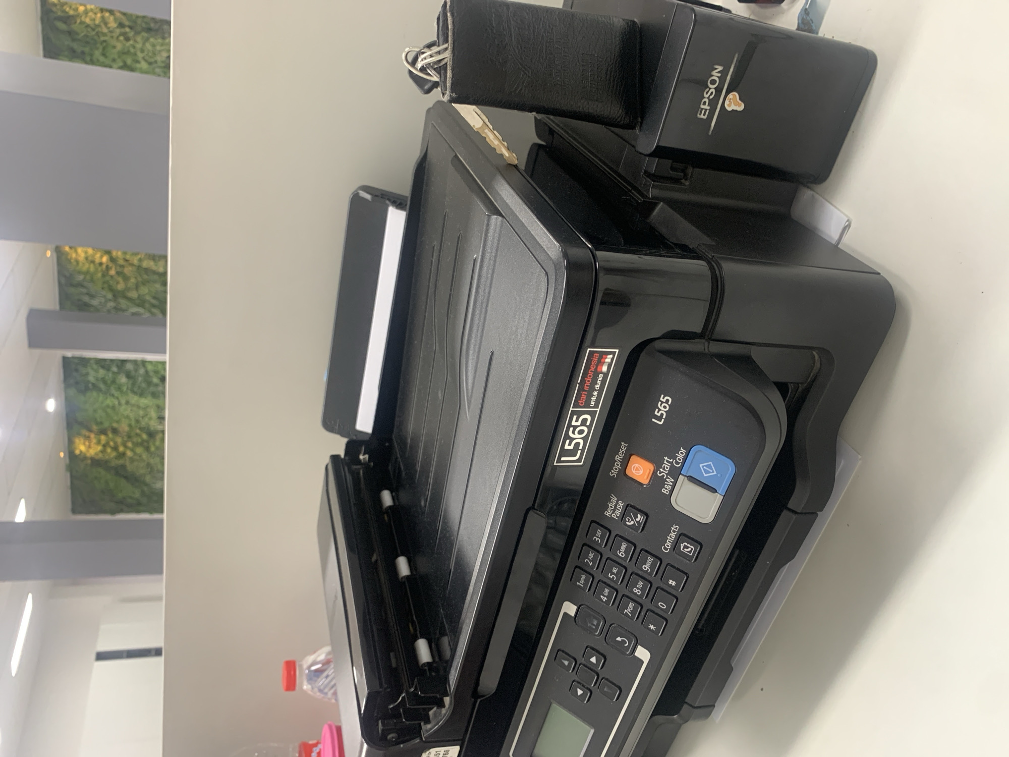 A black Epson L565 multi-function printer is on a white desk in what appears to be an office environment. The printer has a scanner lid on top, a control panel with buttons and a small screen, and an output tray. To the right of the printer, there is a black leather wallet with keys sticking out. The background shows a vertical green plant wall and a gray pillar. The scene is indoors, well-lit, and appears to be a typical office setting. There is no discernible indication of the city of Palembang, Indonesia, or any specific location within it.
