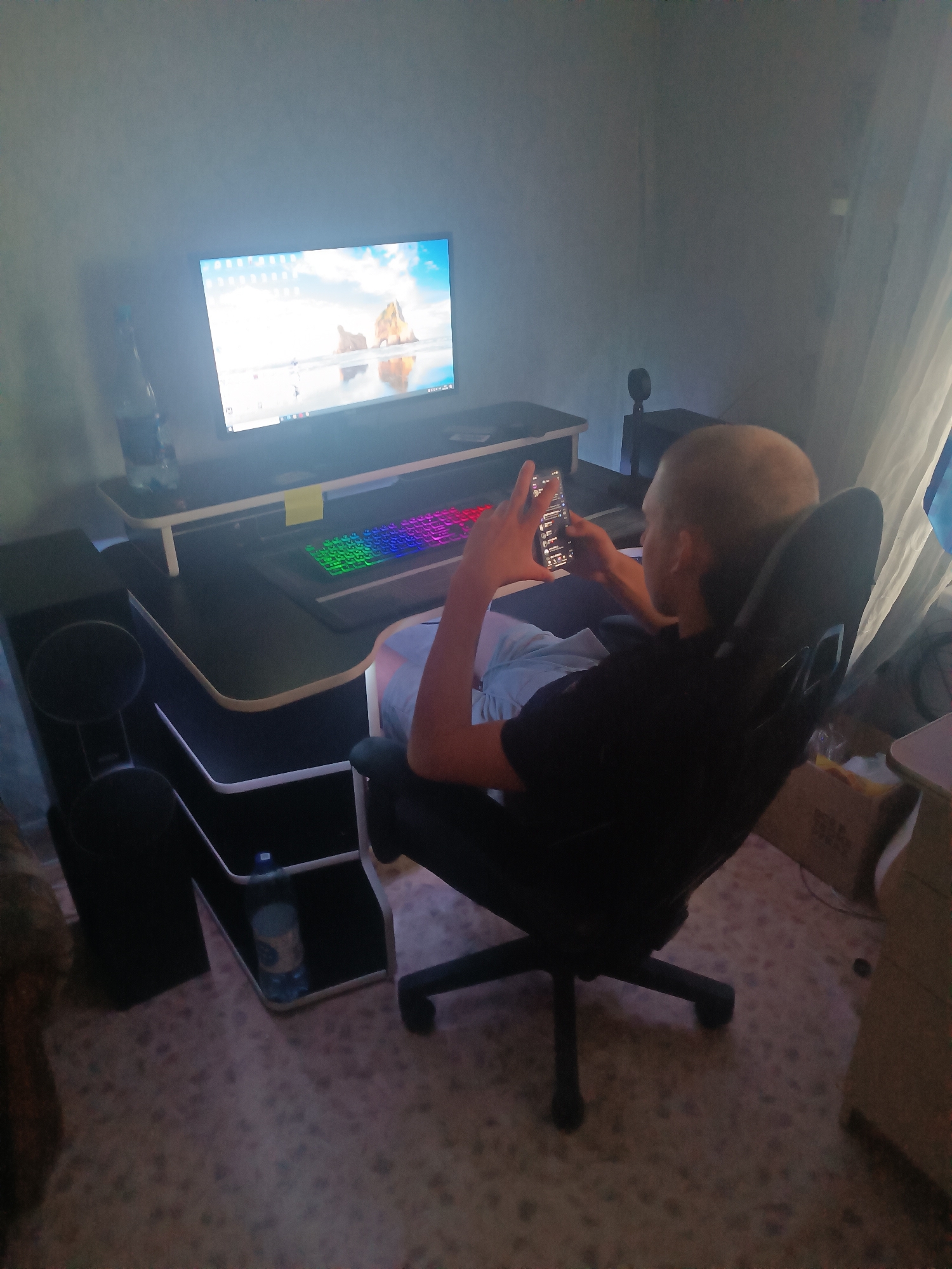 The image depicts a young person, appearing to be male with a shaved head, sitting in a gaming chair in a dimly lit room. The person is holding a smartphone in their hands and appears to be looking at it. In front of the person is a desk with a computer monitor displaying a landscape image, a colorful RGB keyboard, and other accessories. A bottle of water is visible on the desk. To the left of the desk, there is a trash can and a storage unit with shelves. The flooring appears to be carpeted. The background shows a wall and curtains. The scene suggests a typical setup for gaming or computer use. There is no visible indication of the location being in Klyuchi, Russia.