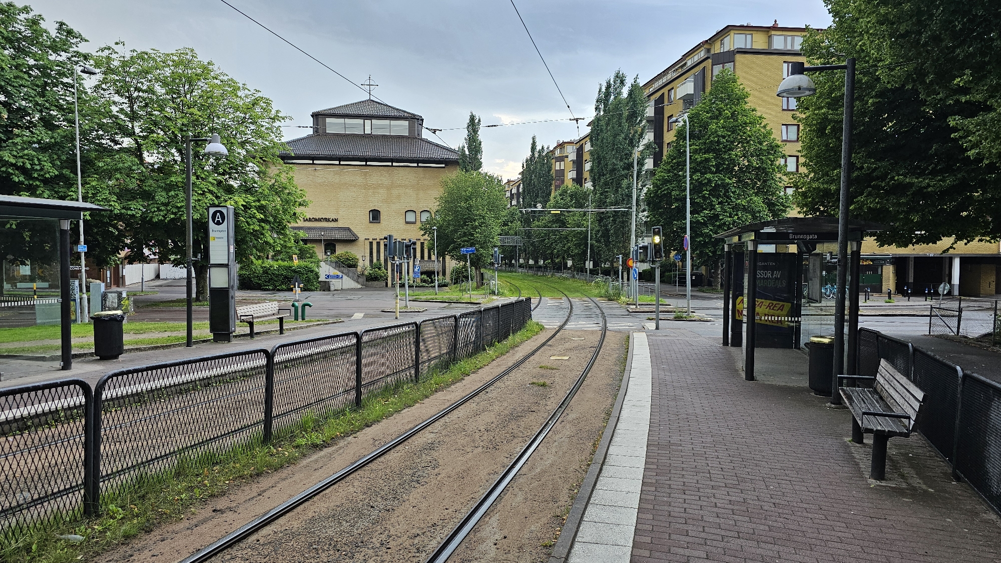 The image depicts a tram stop in Gothenburg, Sweden, specifically at the "Brunnsgatan" stop which serves the "Saronkyrkan" area. In the foreground, tram tracks run parallel to a paved sidewalk and a bus stop shelter. A black metal fence separates the tracks from the sidewalk. A trash can and a wooden bench are visible near the bus stop. The bus stop shelter has advertisements displayed behind glass. Further back, a modern yellow brick building with a cross on its roof, identified as Saronkyrkan (Saron Church), stands prominently. Adjacent to the church is a multi-story yellow apartment building. Green trees and streetlights line the street, and the sky is overcast. The scene is set in an urban environment with no people visible.