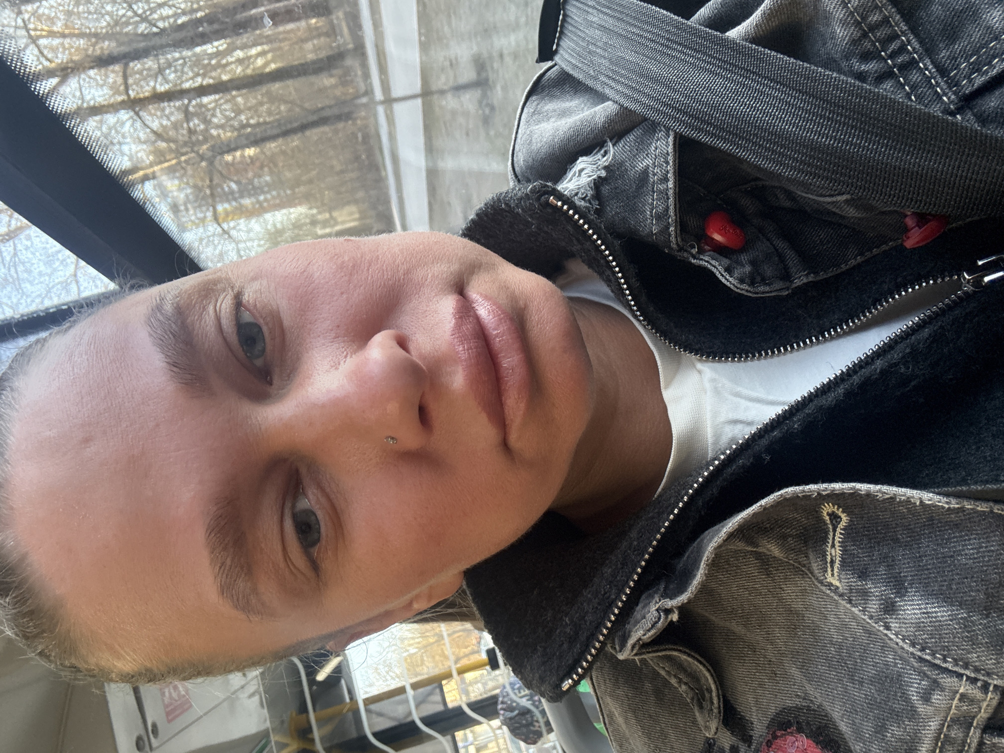A close-up, selfie-style image captures a person with light skin, short, light-colored hair, and blue eyes. The person is wearing a white shirt under a faded black denim jacket with a visible zipper and red button detailing. A backpack strap is also visible. The background shows a window with a view of trees. The person appears to be in transit, possibly on a bus or train. While the image does not reveal the exact location, the information is not readily available to determine if the picture was taken in Perm, Russia.