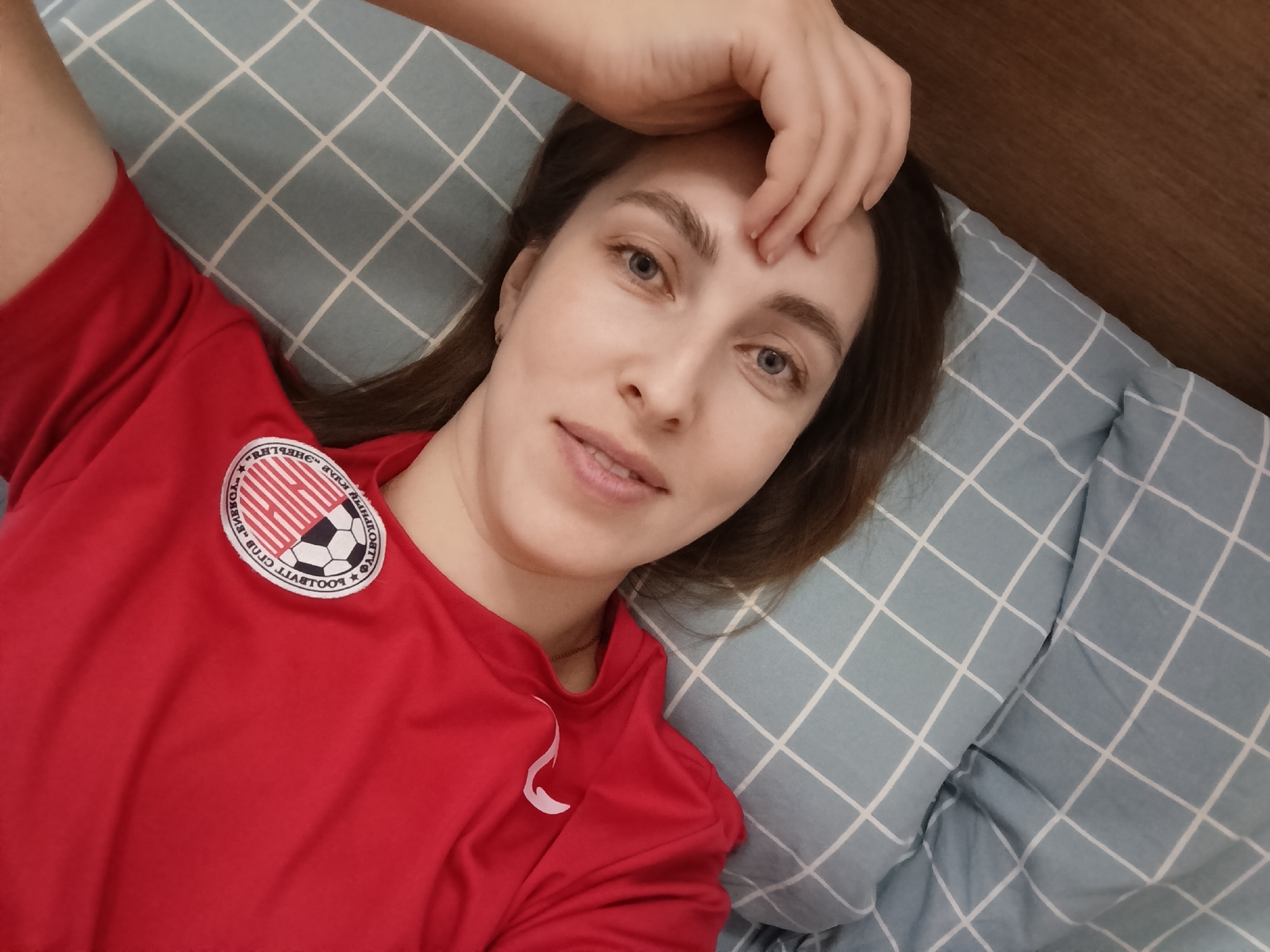 An indoor, eye-level selfie shows a light-skinned young adult female lying on her back, looking directly at the camera with a faint smile. Her long brown hair is visible around her head. Her right hand (viewer's left) rests on her forehead. She wears a red short-sleeved top featuring a circular emblem on the left chest area. The emblem displays a design with red and white stripes, a black and white soccer ball, and mirrored text in Cyrillic and English, indicating "Football Club Energy" and "Chaikovsky". She is positioned on a light blue pillow and bedding with a white grid pattern. A section of dark brown wooden furniture, likely a headboard or wall panel, is visible in the upper right background. The scene depicts an individual in a residential setting within the city of Chaikovsky, Russia.