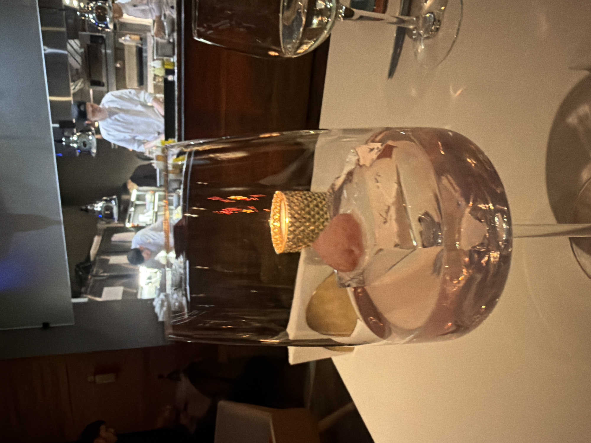 A tilted, medium-shot photograph depicts a culinary scene, likely in a high-end dining establishment. In the foreground, a tall wine glass filled with ice, a small red fruit, and a golden, textured cylindrical object rests on a white tablecloth. The blurred presence of a second wine glass and silverware is seen in the background.
In the mid-ground, a kitchen is visible, populated by chefs in white uniforms. One chef is actively engaged in food preparation.
The specific location in Moscow, Russia is not discernable from the visual information provided.