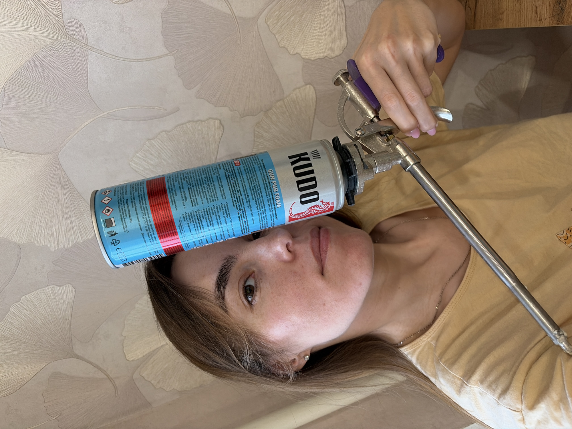 A woman lies on her back with a can of Kudo gun pur foam balanced on her forehead. A metal foam dispensing gun is attached to the can. The can is blue and silver with red stripes and Russian writing. The woman has brown hair and light skin. She is wearing a tan shirt and a necklace. The background is wallpaper with a leaf pattern.