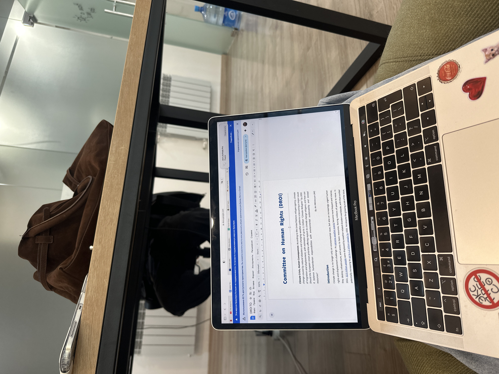 A laptop with a document on "Committee on Human Rights (DROI)" is open on a desk next to a bag. The laptop has stickers on it. The location is likely indoors, potentially in an office, but the specific location in Yerevan, Armenia, is not determinable from the image. There is a water bottle visible in the background and radiator. The subject of the document on the laptop screen pertains to climate change and human rights.
