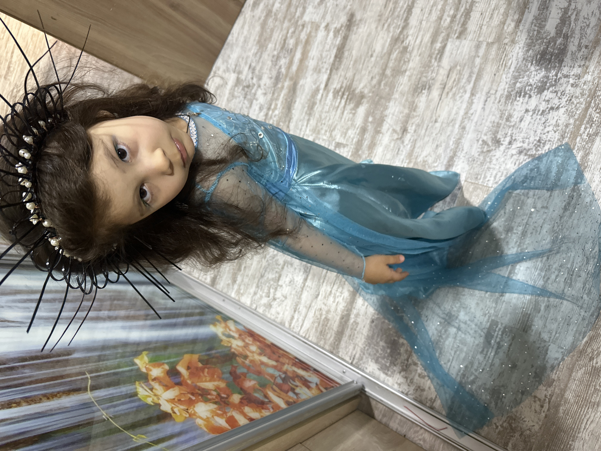 A young child wearing a blue dress and a dark headpiece is angled towards the camera. The background consists of a light-colored, patterned floor. A picture frame is partially visible at the lower left, displaying a warm-toned abstract image. The child's expression is inquisitive.