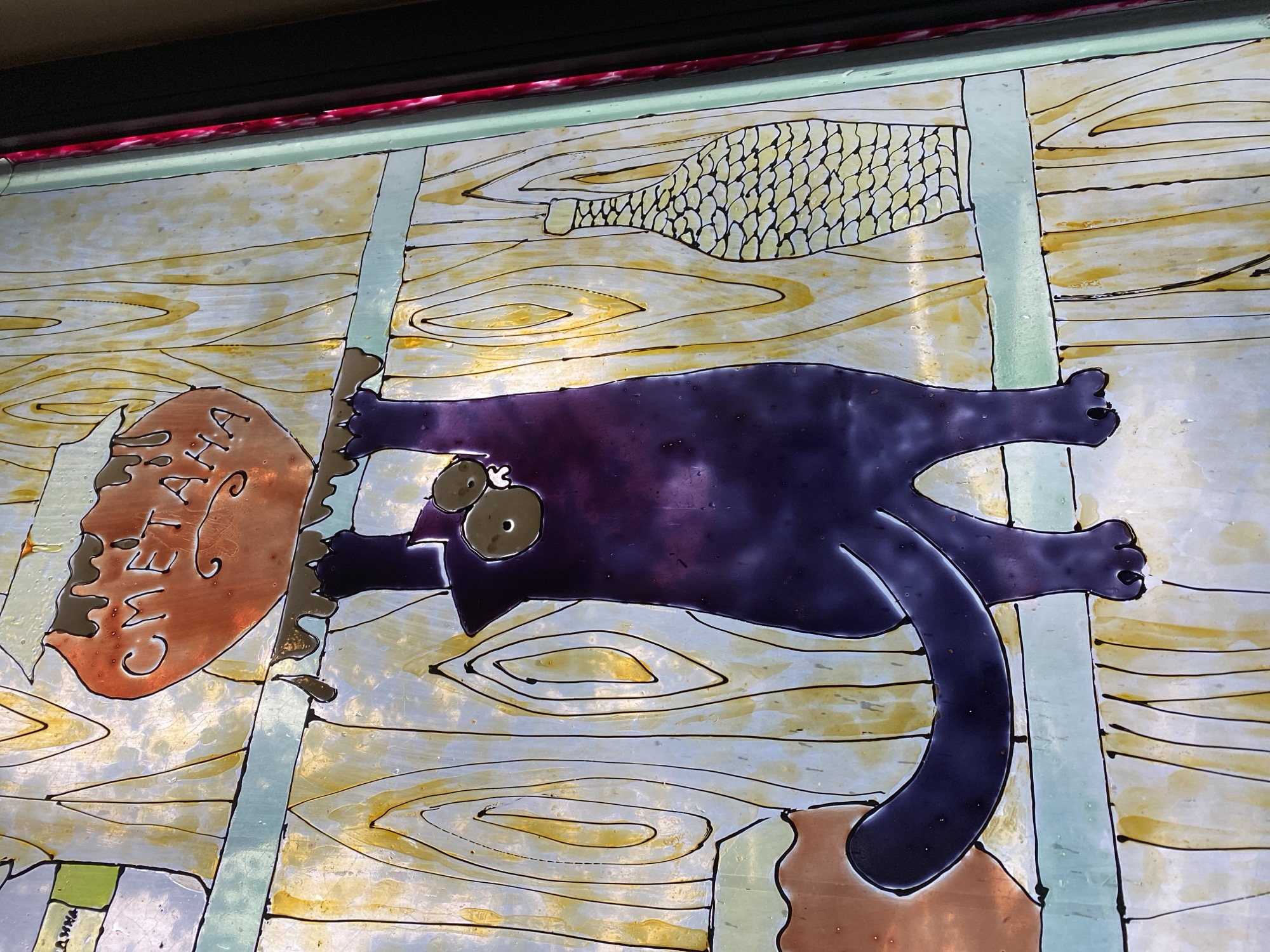 The image displays a close-up, upward-angled view of a stained-glass panel, likely part of a window or decorative screen. The artwork depicts a dark purple cat hanging precariously from a shelf, its paws gripping the edge. Above the cat, a brown, heart-shaped object labeled "СМЕТАНА" (which translates to "sour cream" in Russian and Ukrainian) is also precariously balanced. Drops of what appears to be melted material are shown dripping from both the shelf and the "sour cream" sign.
The background of the stained glass is rendered to resemble wood grain, with vertical lines and swirling patterns in yellow and white hues. To the right of the cat, a stylized, textured bottle with a patterned surface is depicted. To the left and further back, a portion of another container, possibly a jar, is visible. The overall style is whimsical and cartoonish, with bold black outlines defining the figures and objects. The lighting suggests the panel is backlit, illuminating the colors and translucency of the glass.
The specific location in Poltava, Ukraine, cannot be determined from the visual information provided in the image alone. The artwork is the sole subject of the photograph, and there are no contextual clues to pinpoint its exact placement within the city.