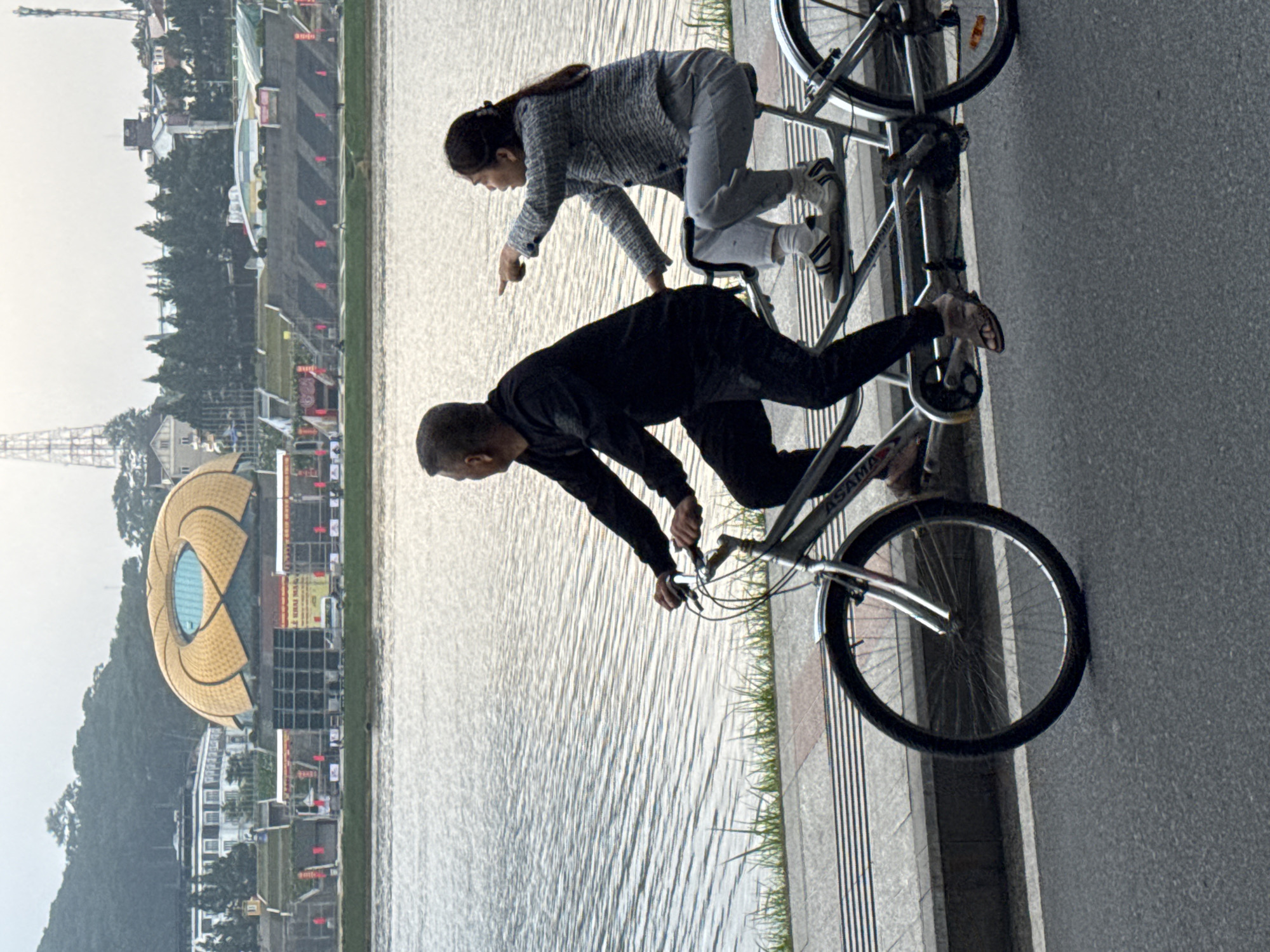 A tandem bicycle with two occupants, a man and a woman, is being ridden along a paved path next to a body of water. The man is in the front, pedaling, and the woman is behind him, pointing towards something off-camera. They are both dressed casually. In the background, across the water, is a city with buildings and hills. One prominent structure resembles a golden, lotus-shaped building. The weather appears to be overcast. The scene is consistent with an outdoor recreational area in a city.