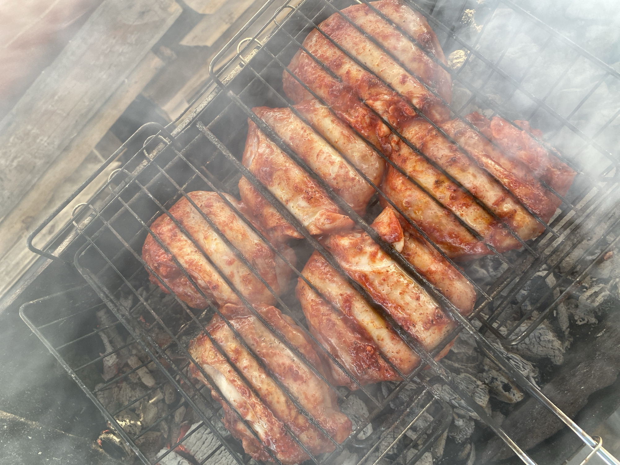 The image shows pork ribs in a metal grilling basket over hot coals, likely being barbecued. There is smoke rising around the food. The background shows some wooden planks. Based on the prompt, this is happening in Chaikovsky, Russia.
