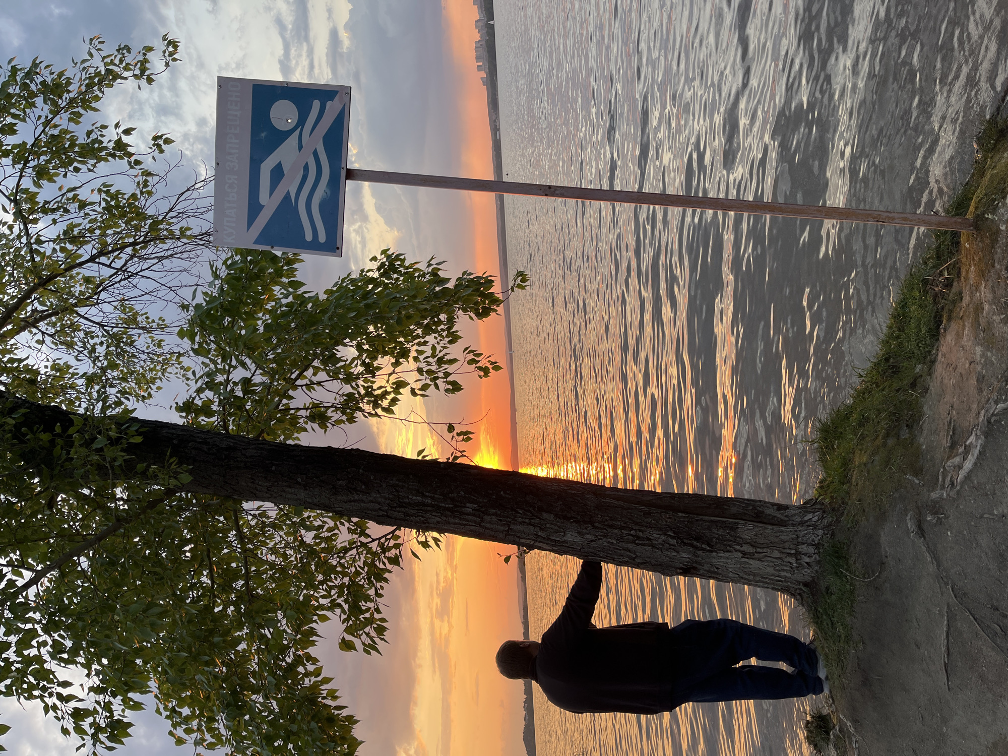 An individual stands on the bank of a body of water during sunset in Yekaterinburg, Russia. The person, wearing a dark jacket and blue jeans, faces the water and appears to be holding onto a tree trunk. Next to the tree is a blue and white sign indicating that swimming is prohibited. The water reflects the colors of the sunset. In the distance, buildings are faintly visible on the horizon. The foreground consists of grass, the tree, and the sign.
