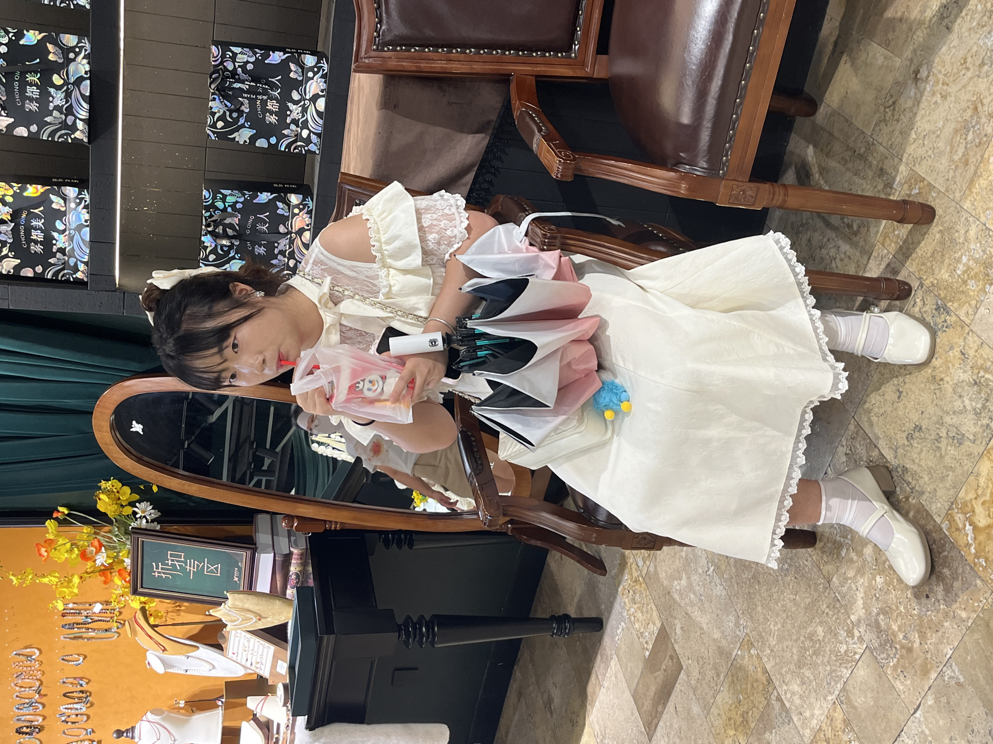 A young East Asian woman sits in a chair, drinking from a pink beverage with a straw. She is wearing a white dress with lace detailing on the sleeves and neckline, white socks, and white Mary Jane shoes. Her hair is pulled back in a bun with a white bow. She holds an open umbrella, which appears to be a set of three umbrellas in pink, white, and black. She is seated in a dimly lit room with ornate wooden chairs and a dark, tiled floor. In the background, there are shelves displaying gift boxes and jewelry. A large, oval mirror with a wooden frame is positioned to the left of the woman, reflecting a portion of the room. The overall setting suggests a retail environment, possibly a boutique or department store.