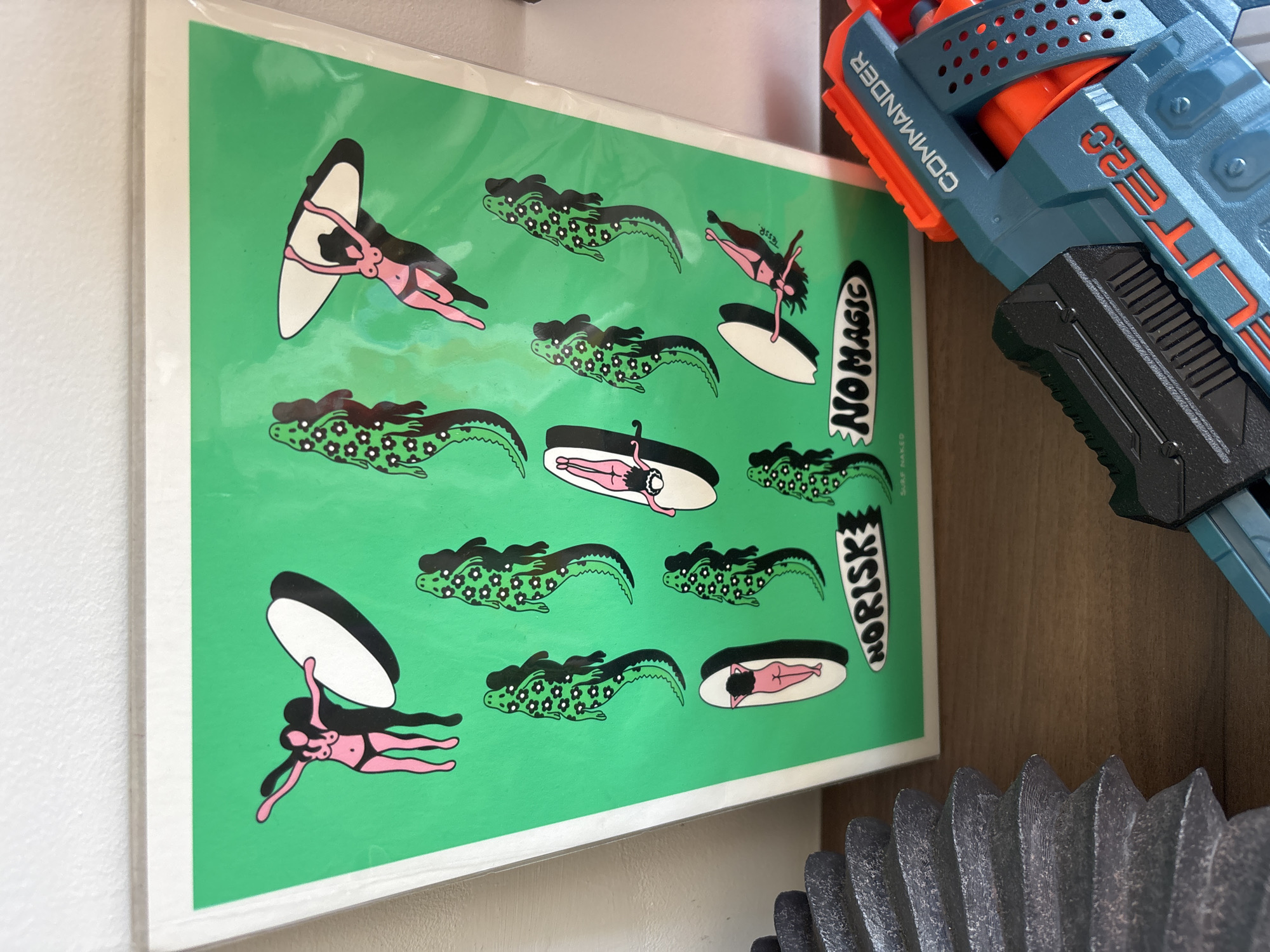 A brightly lit, vibrant green print or poster is showcased indoors, mounted against a white wall. The print features a repeated pattern of stylized illustrations. Several figures of women in pink bikinis are depicted in various dynamic poses, some appearing to float while holding or interacting with white and black surfboards or rings. Interspersed with the women are multiple dark green, alligator-like creatures, each with flowing black human hair and adorned with a pattern of small white flowers.
Two prominent surfboard-shaped elements on the print contain text: one reads "NO MAGIC" vertically, and another says "NO RISK". Smaller, partially visible text includes "ESS.R" near a female figure and "Surf Naked" below the "NO RISK" text.
To the right of the print, a blue and orange toy blaster, identifiable as a Nerf gun, is partially visible. The words "COMMANDER" and "ELITE 2.0" are clearly visible on its side. Below the toy, a dark, textured, and corrugated object, possibly part of an appliance like a fan or speaker, occupies the bottom right corner of the frame. The background consists of a plain white wall on the left and a warm brown wooden surface on the right, indicating an indoor setting, possibly a shelf or desk. The lighting is uniform, suggesting general ambient indoor light. The artwork on the print conveys a whimsical and surreal aesthetic. No specific time of day or other direct location cues for Puebla, Mexico, are visible within the image itself.