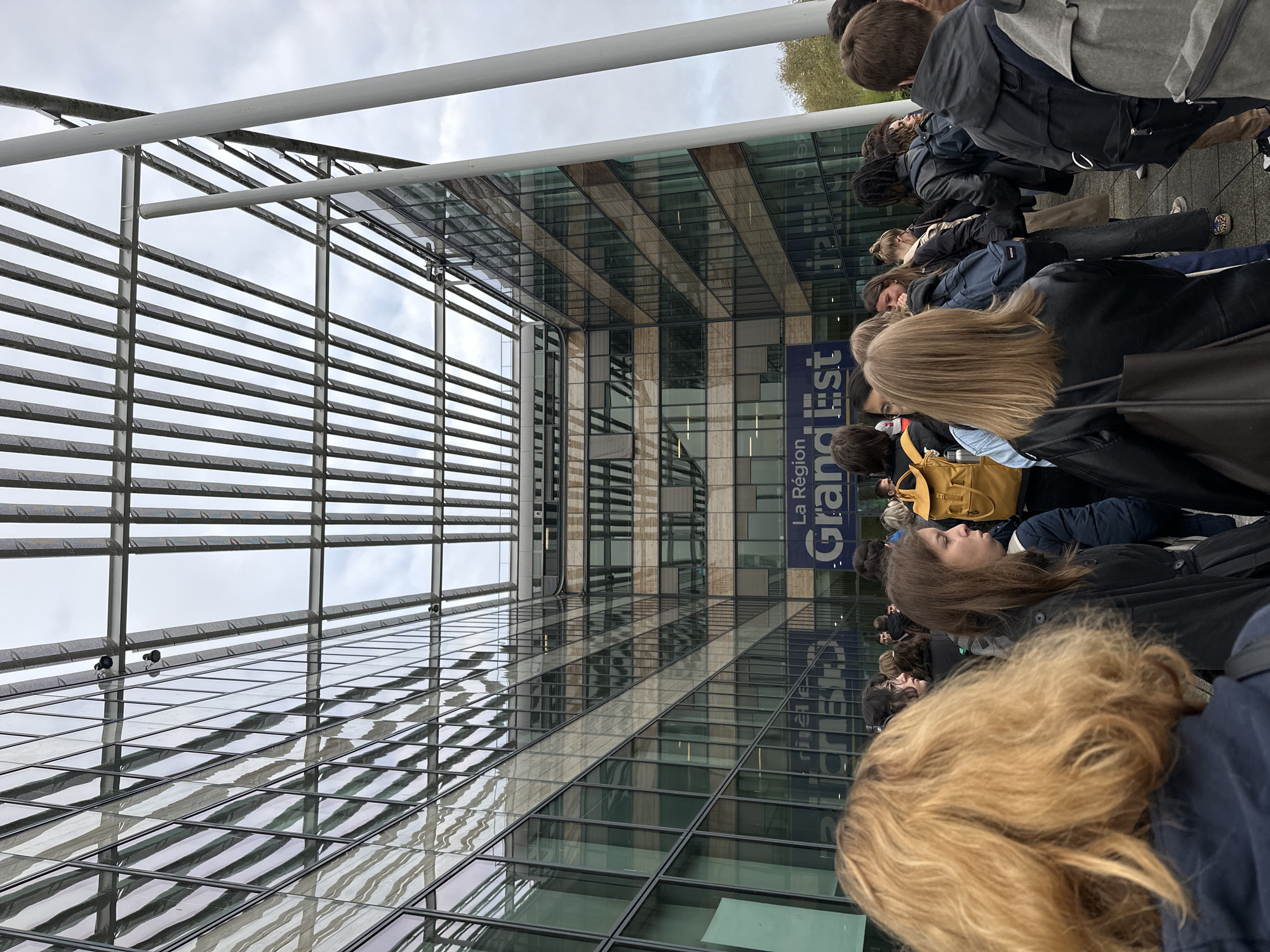 A large group of people is standing in front of a modern building, likely waiting in line. The building features a glass facade and a prominent sign reading "La Région Grand Est," indicating the location is Strasbourg, France. The exterior of the building is constructed with metal grates and panels, creating a geometric pattern. The crowd consists of people of various ages and appearances, some with backpacks, suggesting they may be students or visitors. The sky is overcast, indicating a cloudy day.