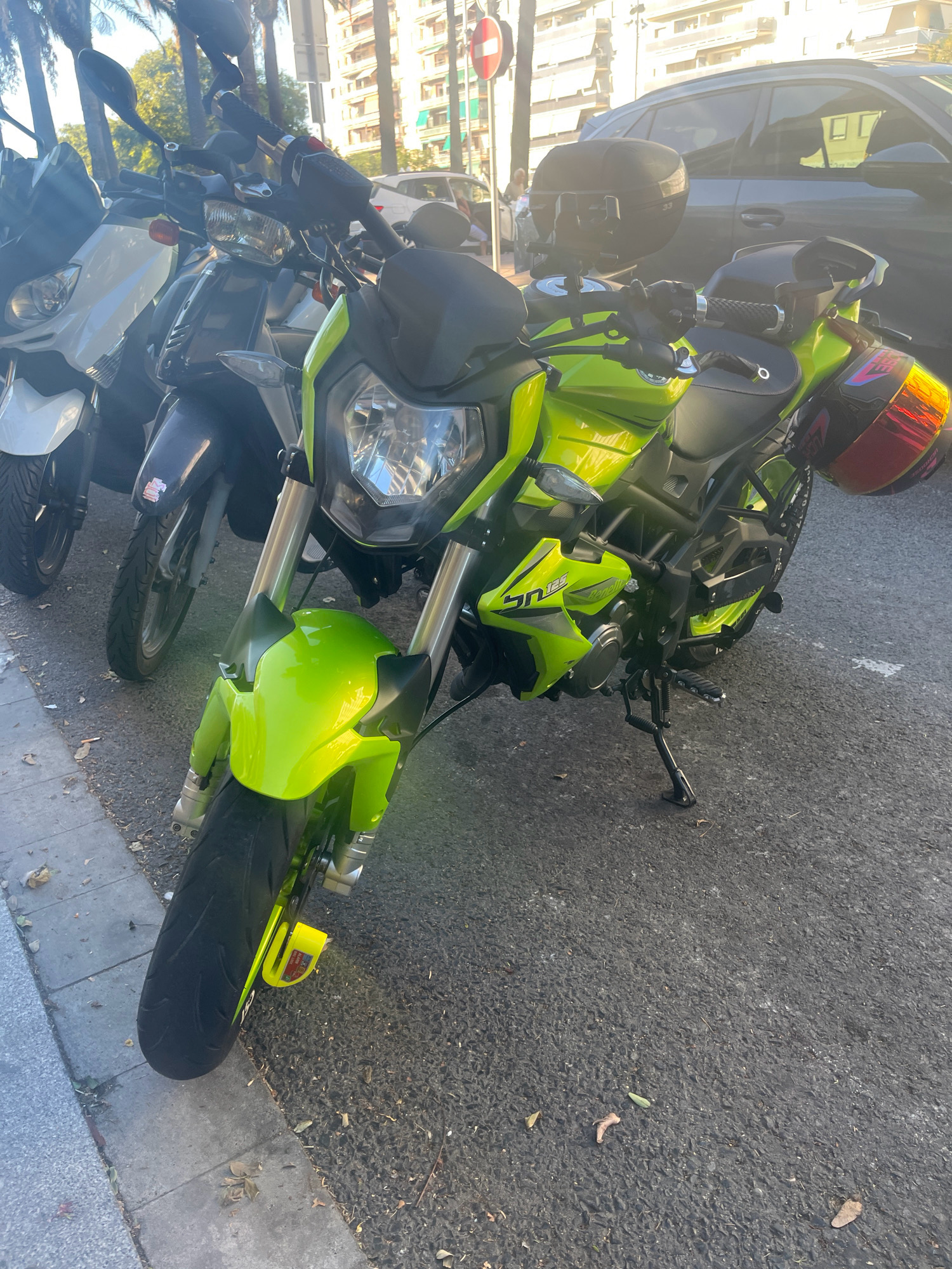 A neon green Benelli SN 125 motorcycle is parked on a city street in Tarragona, Spain, alongside other parked scooters. The motorcycle is the primary focus, positioned at a slight angle facing forward. It features a prominent headlight, handlebars with mirrors, a black seat, and a red and yellow helmet secured to the rear. The motorcycle's frame and wheels are also neon green. In the background, a dark-colored SUV is parked, and beyond that, buildings with visible windows and balconies line the street. Palm trees and other greenery are present, suggesting a warm climate. The ground is paved with asphalt, and a concrete curb is visible to the left, with some fallen leaves scattered on the ground. The scene is illuminated by natural sunlight, casting shadows. There are no people clearly visible in the immediate vicinity of the motorcycles.