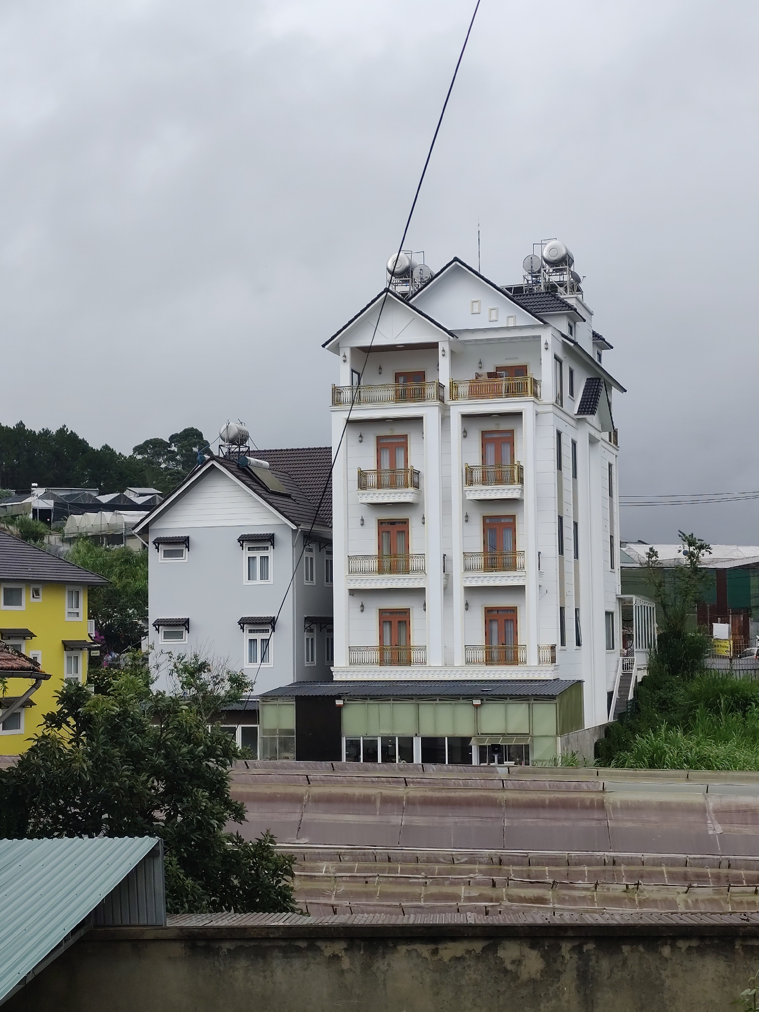 The image captures a view of a modern white hotel building with multiple balconies, located in the city of Lang Biang - Đà Lạt, Vietnam. Adjacent to the hotel is a light gray residential building with a dark roof and several windows. A bright yellow building is visible further to the left. The scene is set against a backdrop of lush green hills and trees, with a slightly overcast sky. In the foreground, there is a rooftop with corrugated metal sheeting and a concrete wall. The overall impression is that of a densely built urban or semi-urban area within a hilly landscape.