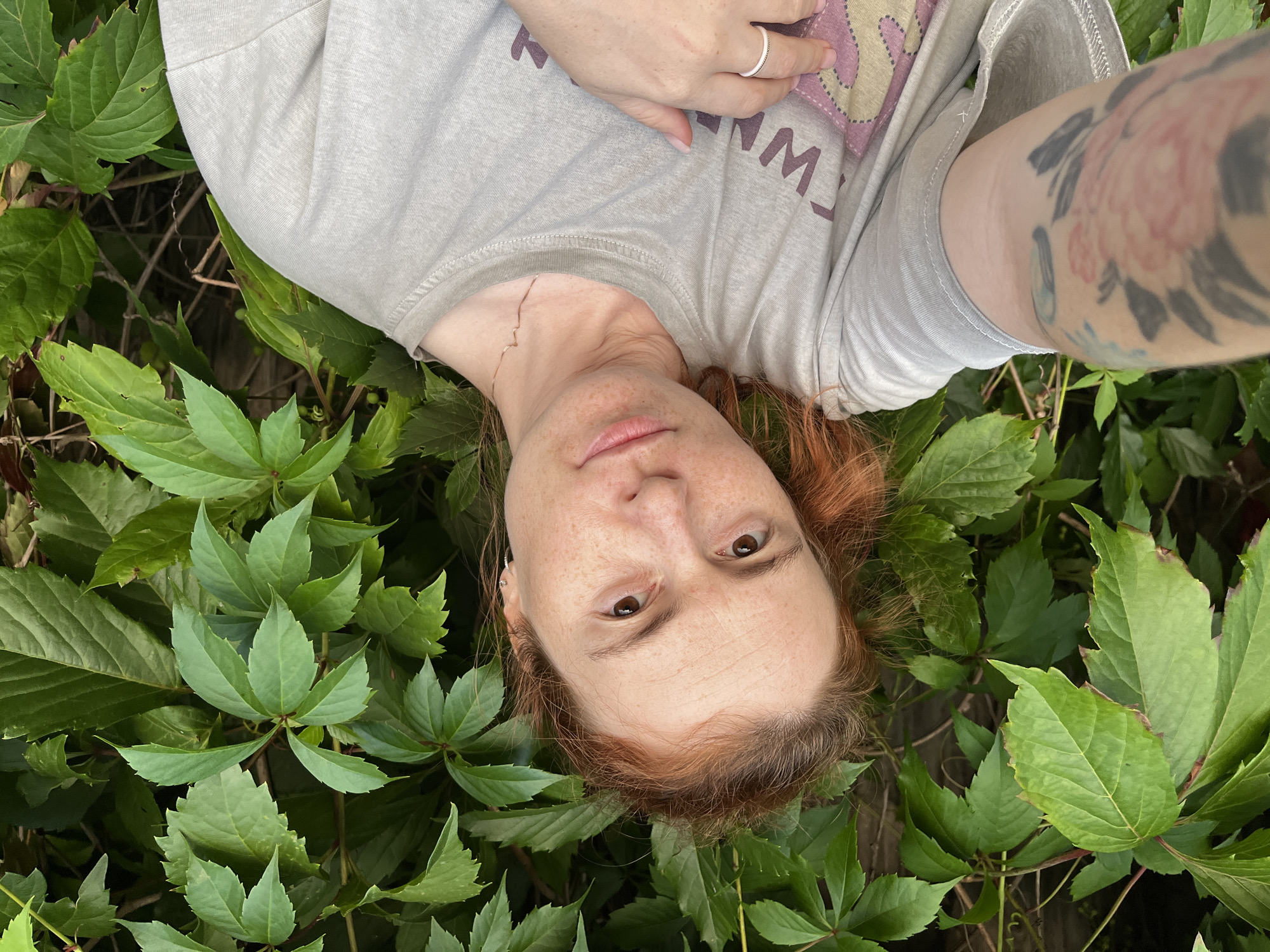 A young woman with reddish-brown hair and freckles is taking a selfie outdoors in front of a dense wall of green leaves. She is wearing a light grey t-shirt. Her left arm, with a visible tattoo of a pink flower, is extended as she holds the camera. Her right hand rests on her chest, wearing a silver ring on her ring finger. The background consists of lush, green foliage with some wooden fence visible through the leaves. The lighting suggests it is daytime. The image does not provide specific details about the city of Igra, Russia, beyond the implied outdoor setting.