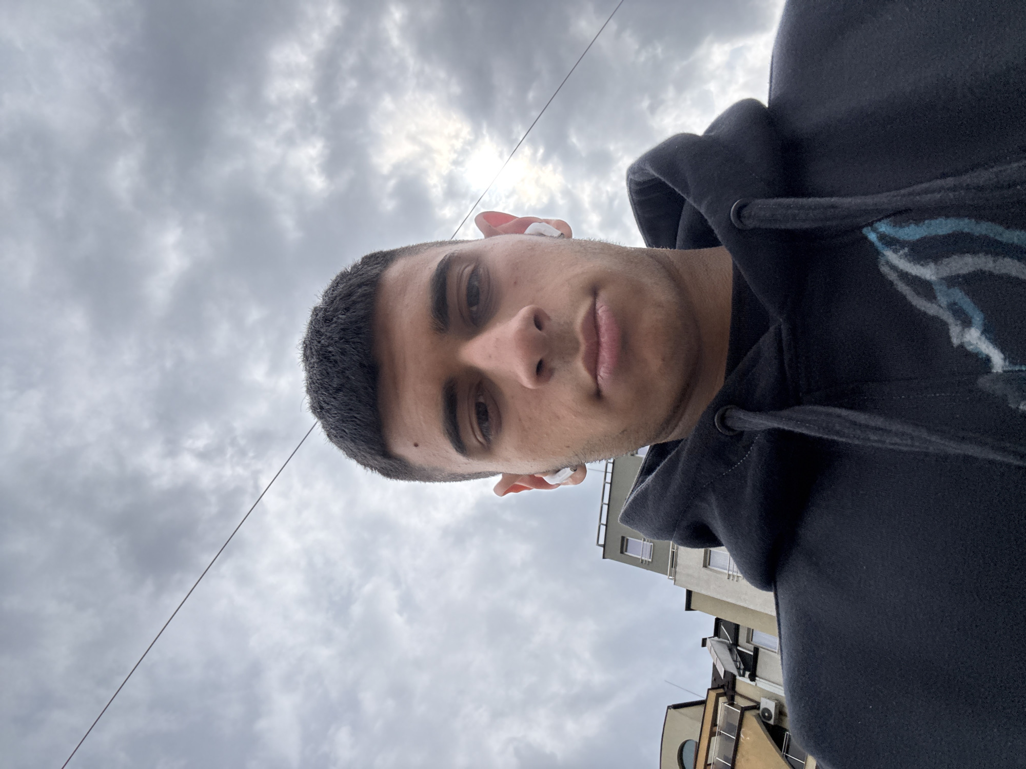 The primary image is rotated 90 degrees counter-clockwise. When viewed upright, it features a head-and-shoulders shot of a young man, looking directly at the camera from a low-angle perspective.
**Subjects:** The main subject is a young man, likely in his late teens or twenties, with short dark hair and a clean-shaven face. He has a neutral, calm expression. White earbuds are visible in both ears. He is wearing a dark, possibly black, hooded sweatshirt, which features a light blue and white abstract or graphic design on the right shoulder/chest area.
**Scene/Setting:** The background is predominantly an overcast sky with patches of brighter light, suggesting the sun is behind the clouds. A thin wire or cable stretches diagonally across the upper portion of the sky. In the lower right area of the image, parts of several urban buildings are visible, including windows, balconies, and an air conditioning unit on one of the structures.
**Activity or event taking place:** The man is being photographed, likely in a selfie or a shot taken from a very low angle, with his face centered against the sky and buildings.
**Notable details:** The weather is cloudy and overcast. The time of day appears to be daytime. The urban buildings in the background provide a location cue consistent with a city setting, specifically Sofia, Bulgaria, as provided. There is no visible text in the image.