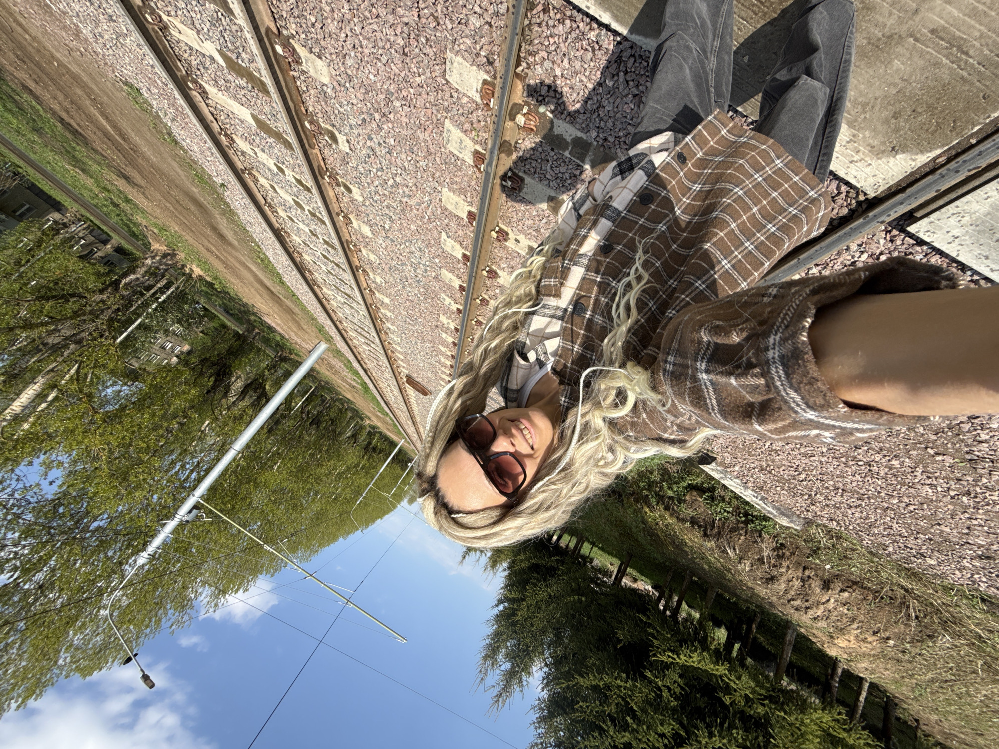 A selfie is taken outdoors in Shebunino, Russia. A person with long, blonde, curly hair, wearing sunglasses, and a brown and white plaid shirt is positioned in the lower right foreground. The subject is smiling and looking towards the camera.
The background consists of railway tracks running diagonally from the upper left to the lower right. The tracks are lined with gravel. To the left of the tracks is a grassy area and trees under a blue sky with scattered clouds. To the right, next to the tracks, is a concrete platform and additional vegetation. Utility poles and lines are visible on the left side of the image.