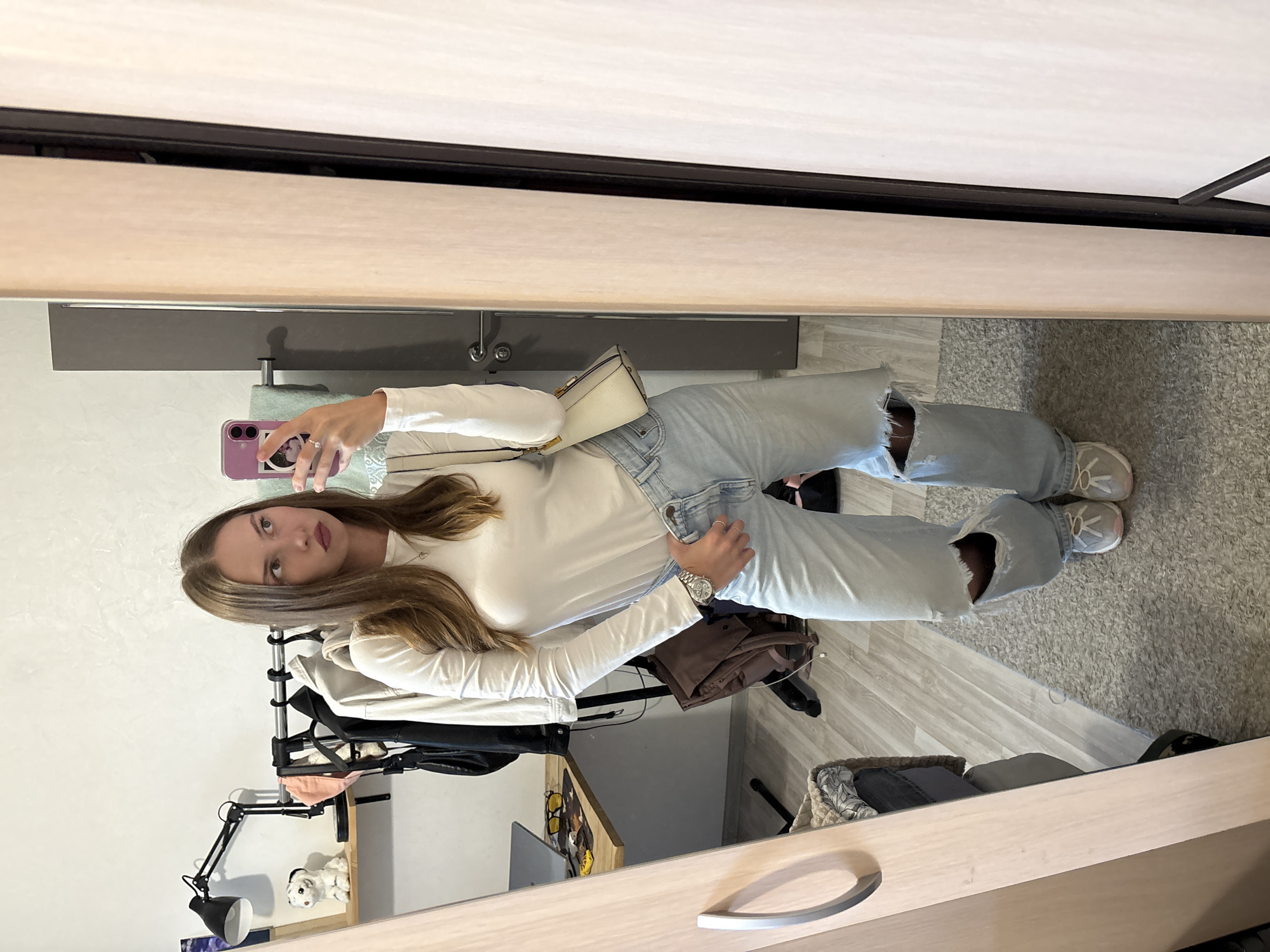 A young woman is taking a mirror selfie in what appears to be a bedroom. She is wearing a white long-sleeved shirt, light-wash ripped jeans, and light-colored sneakers. She has a small, light-colored handbag slung over her shoulder. A phone is held up to take the picture. In the background, there is a clothing rack with several items of clothing, a desk with a laptop and some accessories, a shelf with a decorative item, and a mirror. The floor is covered with a gray rug and wooden flooring. The overall impression is that of a casual, indoor setting. The metadata indicates the photo was taken in Perm, Russia.