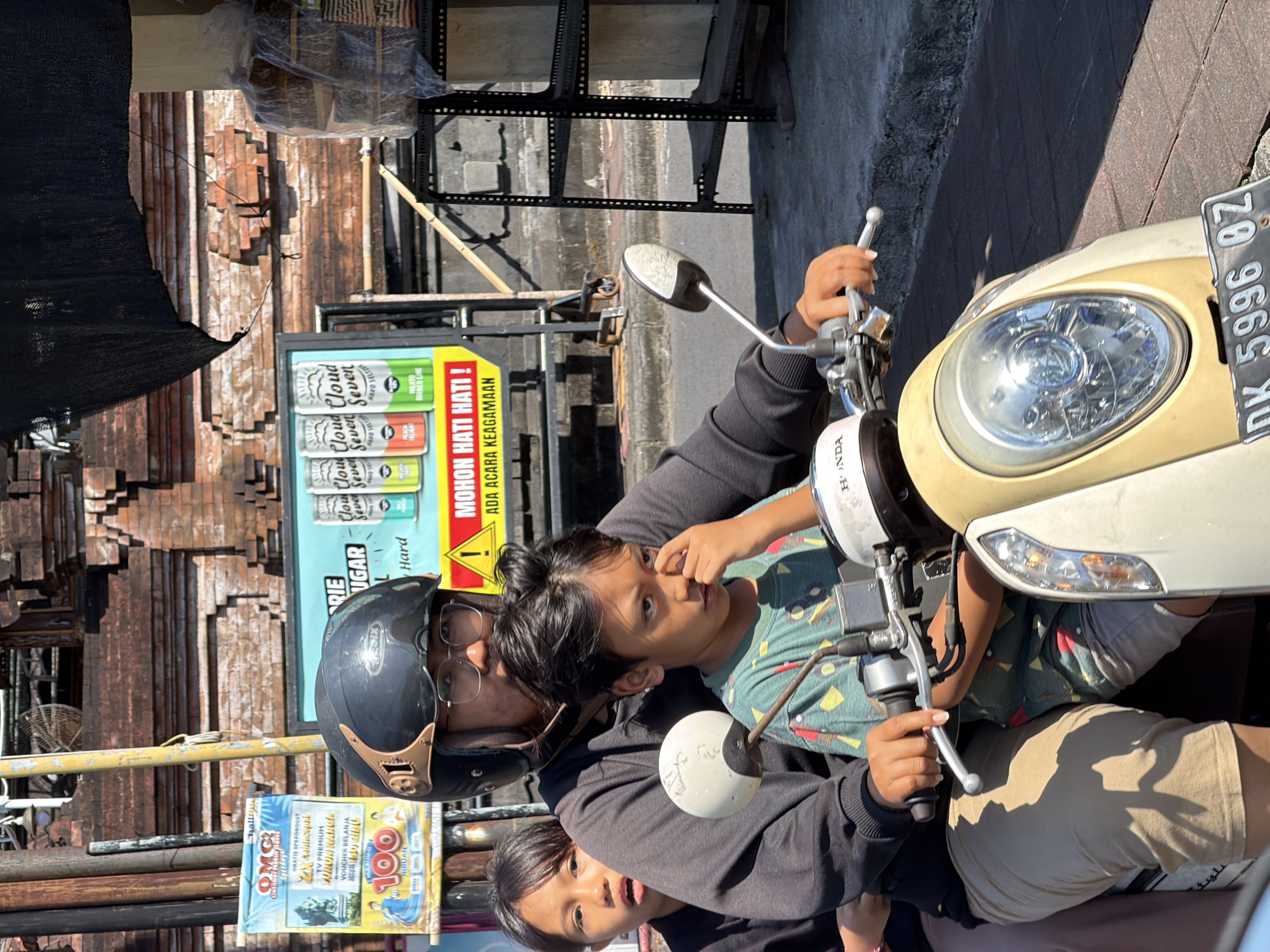 In Kuta Selatan, Indonesia, on a sunny day, a man wearing a black helmet and glasses is riding a scooter, carrying two children. The man is in the driver's seat, with a younger boy in front of him, and a girl behind him. The boy is wearing a green and yellow patterned shirt and has his hand to his nose. The girl is wearing a black long-sleeved shirt and has an open-mouthed expression. They are on a paved street, with a brick building in the background. To the left of the scooter is a poster with text and images, and behind the scooter is a rack of goods covered in plastic. The scooter is light-colored and has a license plate that reads "DK 5996 BZ". The overall scene depicts a family traveling together on a scooter in an urban environment.