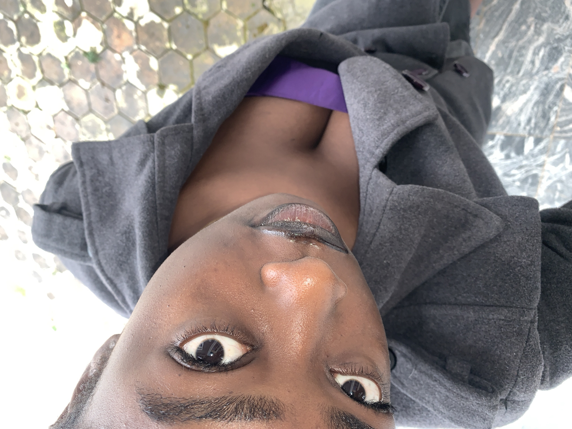The image is a close-up, high-angle shot of a young Black woman from the chest up. She is wearing a dark gray coat over a vibrant purple top. Her face is the primary focus, with her eyes looking directly at the camera. Her skin is dark and appears to have a dewy sheen. Her lips are full and appear to be coated with a dark, glossy substance. Her eyebrows are dark and well-defined, and she has long, dark eyelashes.
The background is out of focus but shows what appears to be a paved outdoor area with hexagonal tiles. There is some greenery visible in the far background. The lighting suggests it is daytime. The scene does not provide specific visual cues to confirm the location as Gura Top, Nigeria.