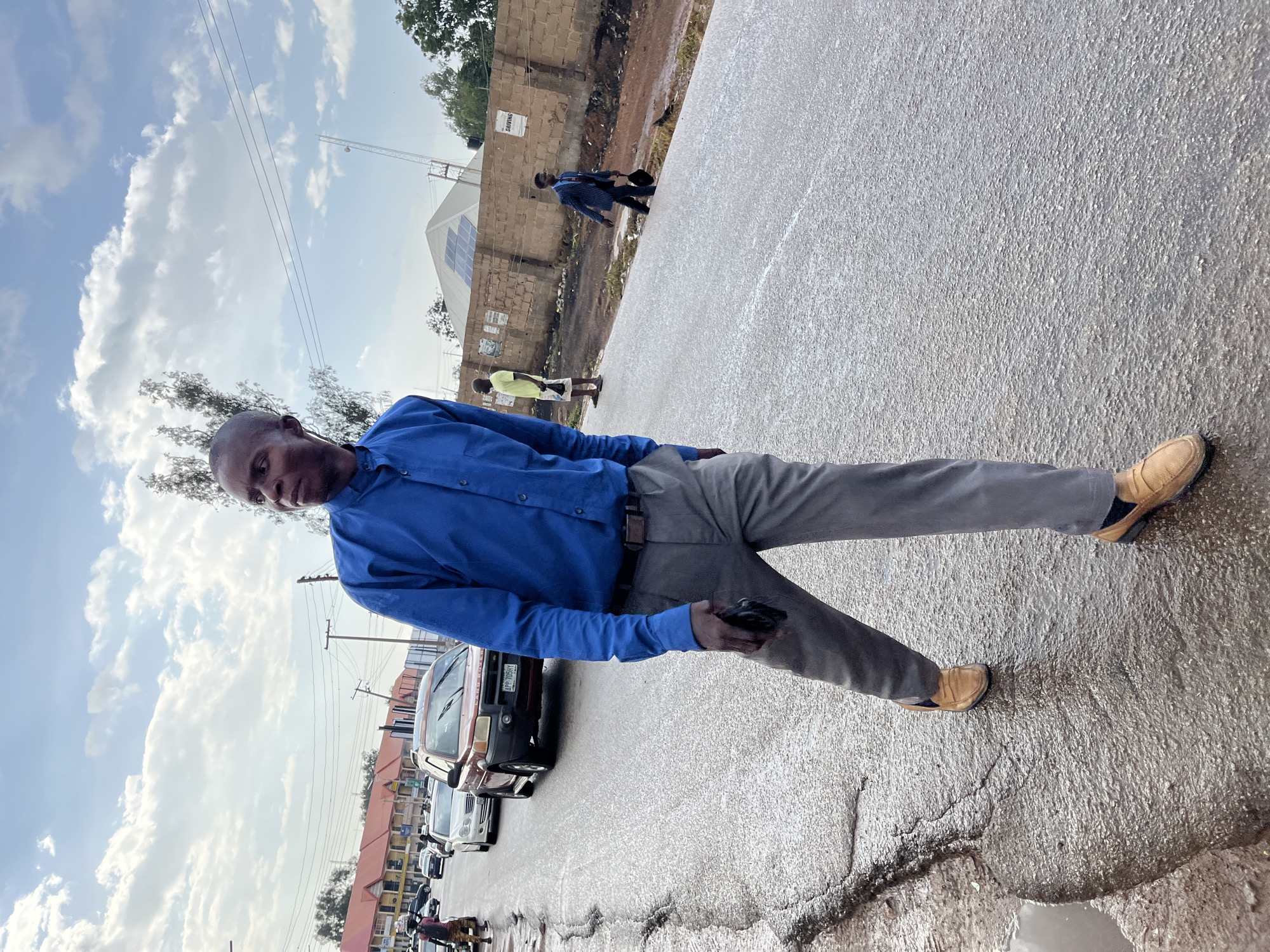 The image captures a full-length shot of a man posing on a wet, paved road in Gura Top, Nigeria, under a partly cloudy sky. The man, positioned centrally, is wearing a blue long-sleeved collared shirt, grey trousers, and light brown slip-on shoes. He is bald, and his gaze is directed towards the camera. He holds a dark object, likely a mobile phone, in his left hand, which is resting at his side.
The road surface appears rough, with visible cracks and puddles, suggesting recent rain. To the left of the man, several vehicles, including what looks like a red van (partially visible license plate "AFP-06X..."), are parked along the roadside. Beyond the vehicles, various buildings, including multi-story structures with orange roofs, line the street. Utility poles with overhead wires are also visible.
In the background, on the right side, there's a low wall constructed from reddish bricks. On this wall, a light-colored sign with text, possibly "GAWUNA," is partially visible. Behind the wall, further back, a large white structure with solar panels on its roof stands out. Several other individuals are scattered in the background; one person in a blue shirt is walking near the wall, and another in a light yellow top is standing further down the road. The overall setting is a bustling, semi-urban street scene during the daytime, with clear, bright conditions despite the cloudy sky.