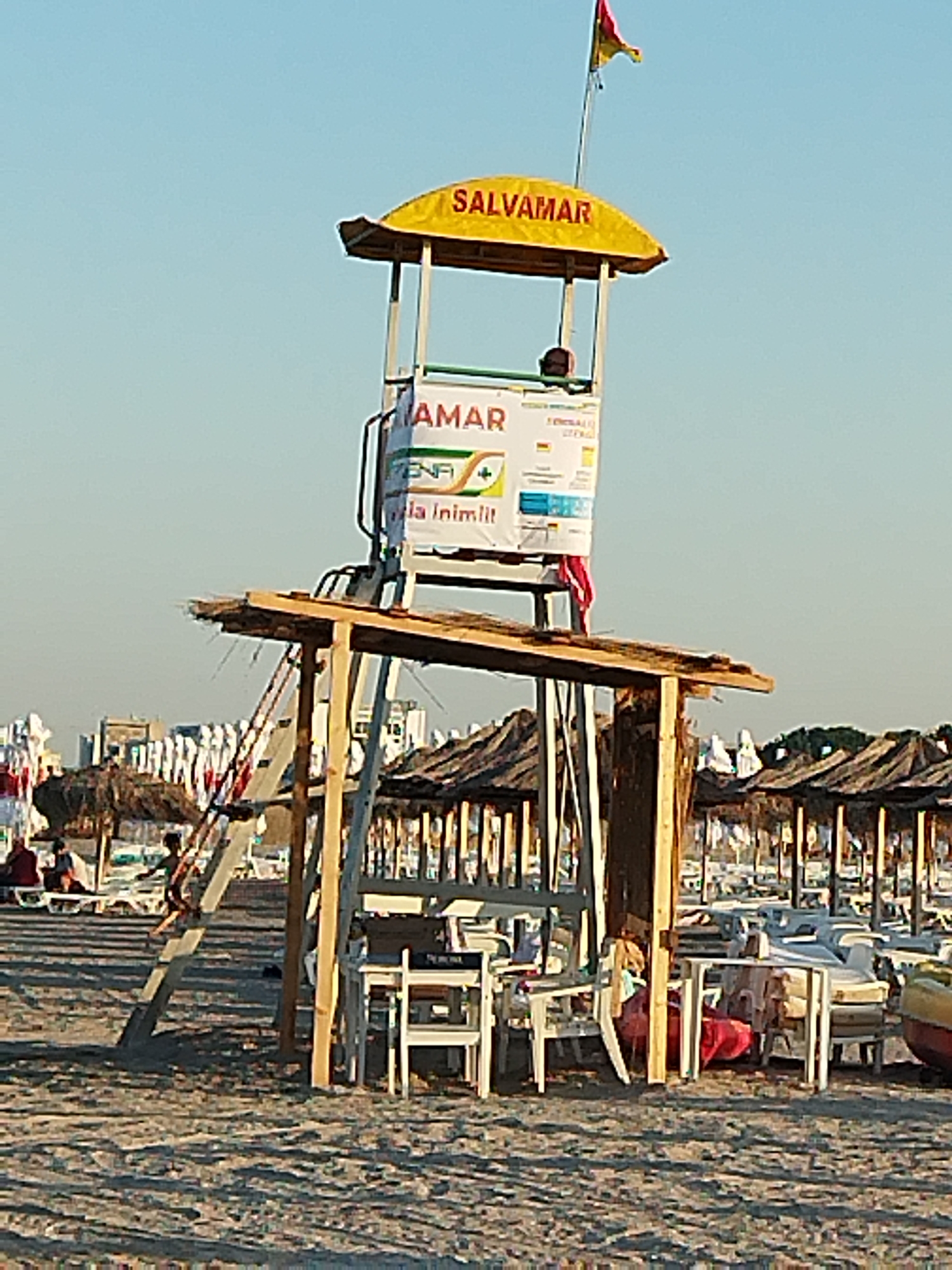 The image shows a lifeguard tower on a beach. The tower is yellow and has the word "SALVAMAR" written on it. There is a flag at the top of the tower. Below the tower, there are chairs and tables. In the background, there are beach umbrellas and people. The scene appears to be on a sunny day. The location is the beach of Constanța, Romania, a resort city on the Black Sea coast known for its beaches and summer tourism.
