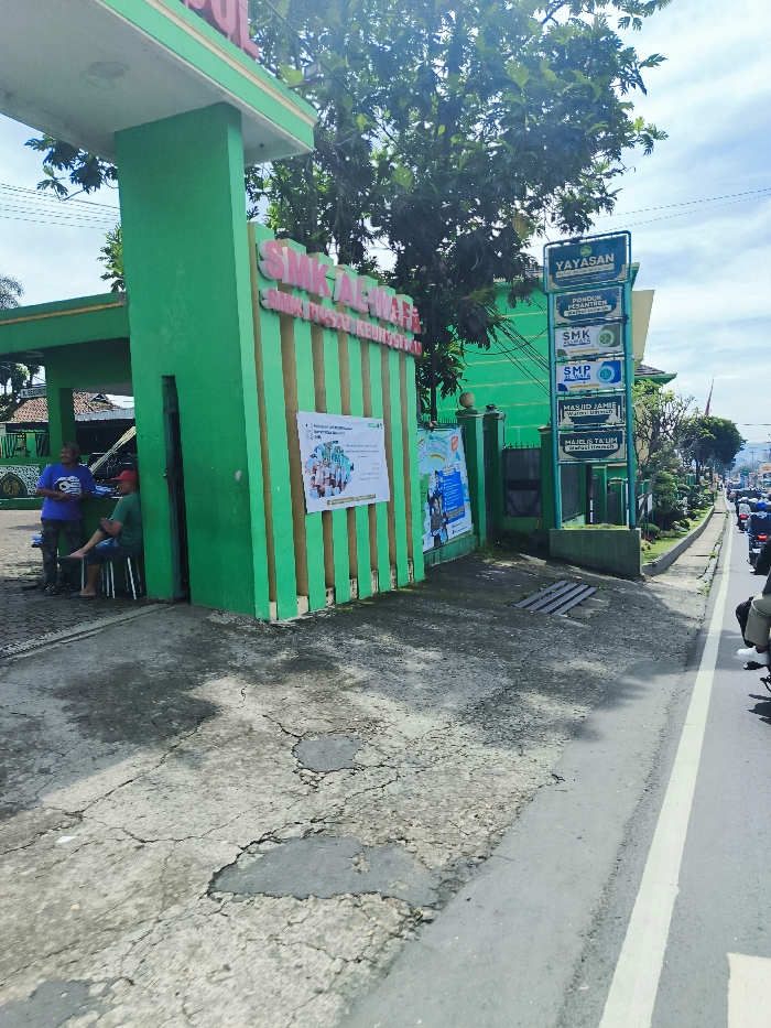 The image shows the entrance to the SMK Al-Wafi, a school located in Ciwidey, Indonesia, at coordinates -7.107 latitude and 107.455 longitude. The entrance is marked by a large green gate with the school's name and "SMA/SMK/SMP" written on it. There are two men standing near the gate, one wearing a blue shirt and the other wearing a red shirt. A sign on the right-hand side of the gate advertises "Yayasan Pondok Pesantren Al-Wafi", a religious school. The sidewalk is made of cracked concrete. A road runs along the right side of the image, and a few motorcycles can be seen driving on it. The background features a green tree, green vegetation, and a clear blue sky.